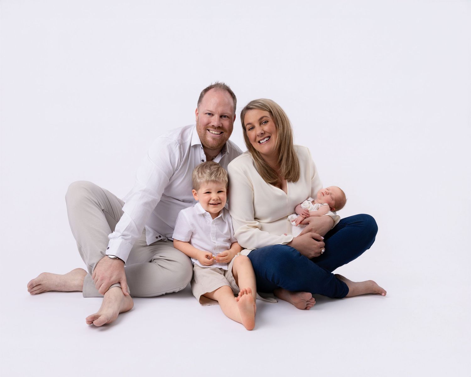 Happy family of four poses on white background, parents smiling with toddler and newborn baby in a warm portrait.