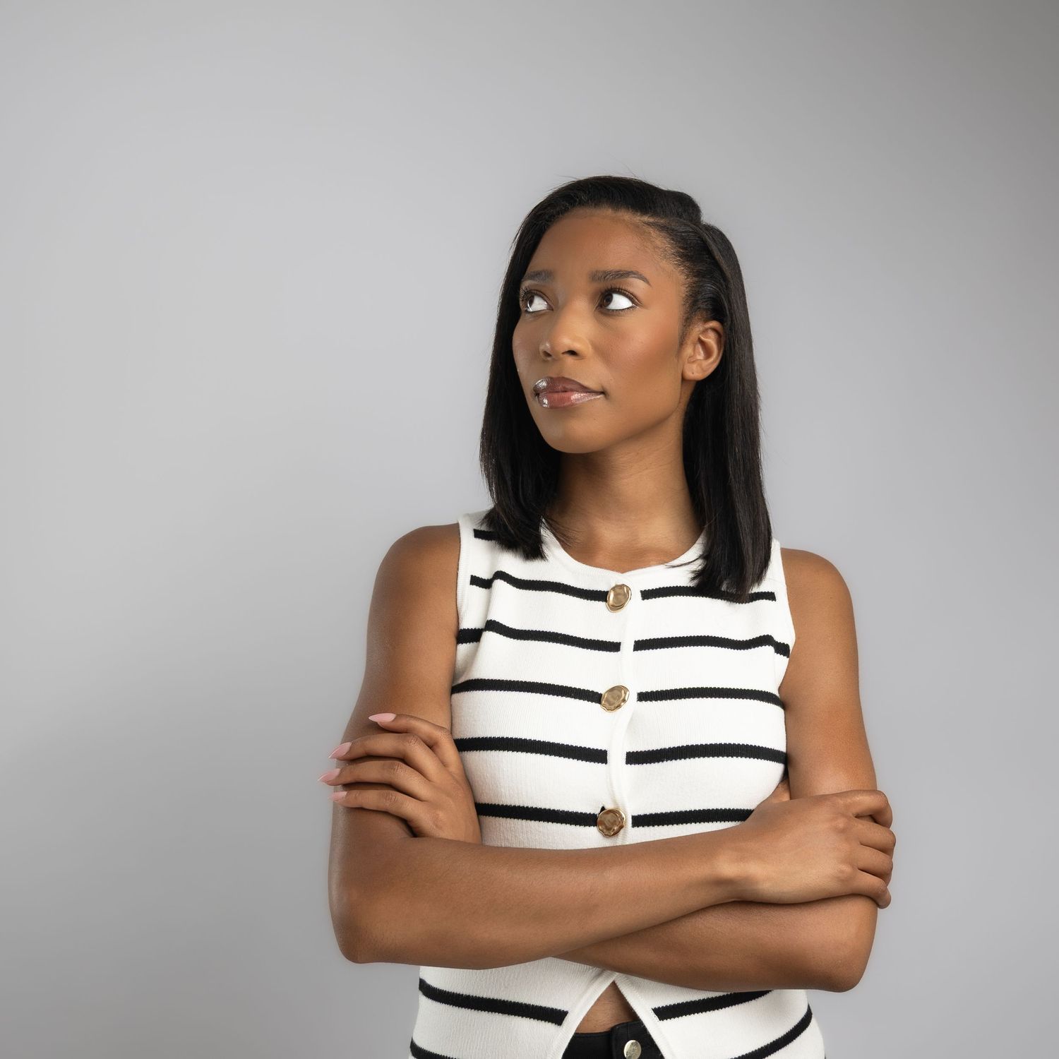 Young Black woman in striped sleeveless top stands arms crossed, glancing sideways, against gray background.
