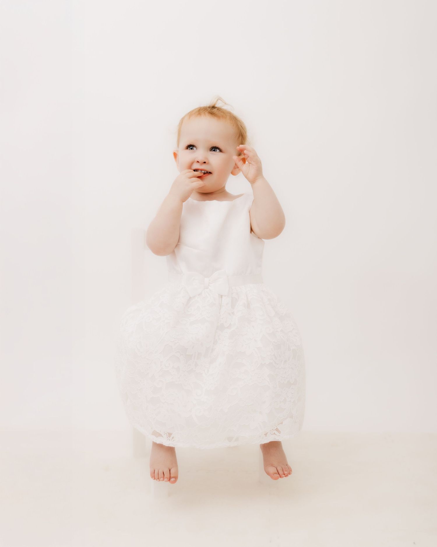 Baby in white lace dress sitting and playing against white background.
