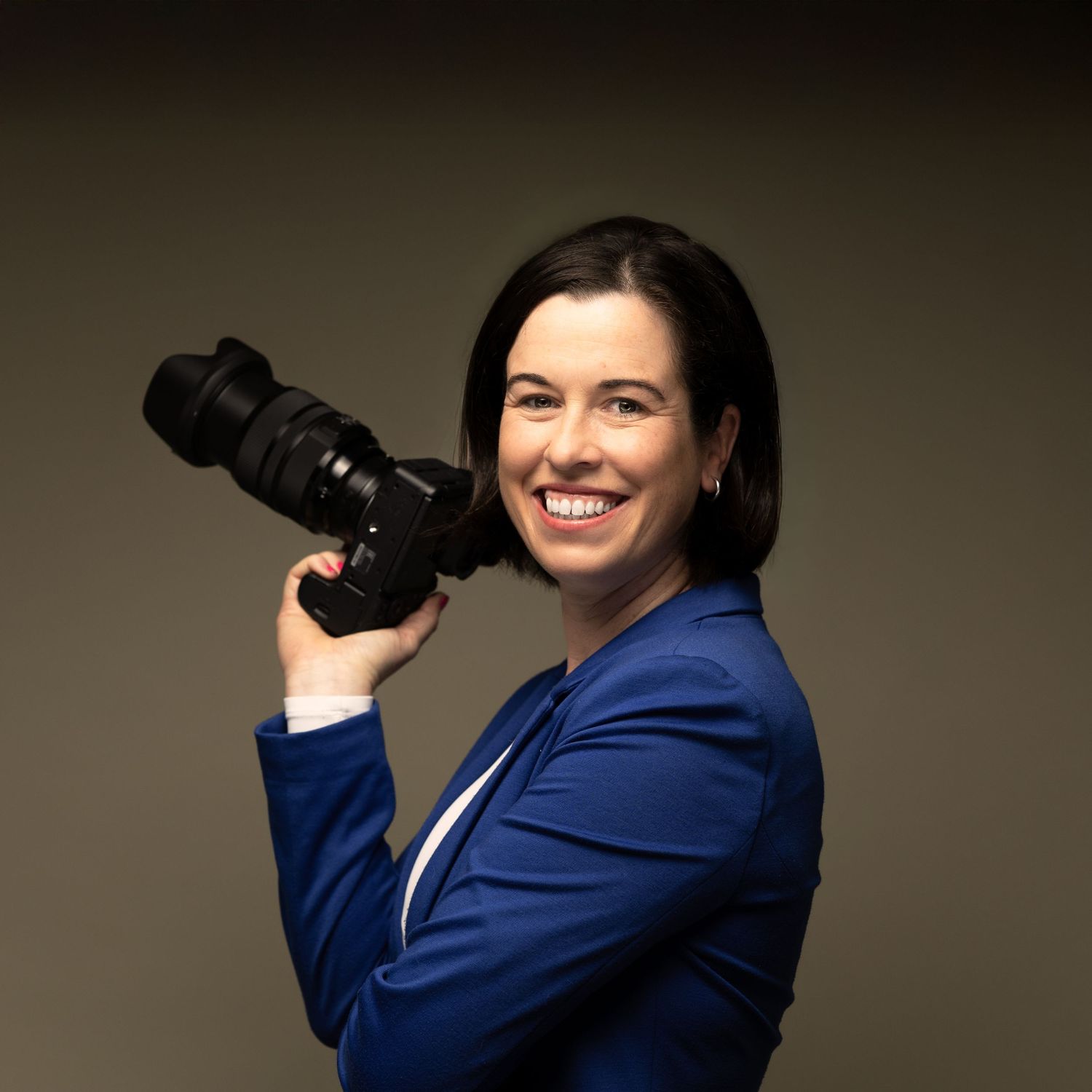 Professional photographer in blue blazer demonstrates different camera poses against neutral studio background.