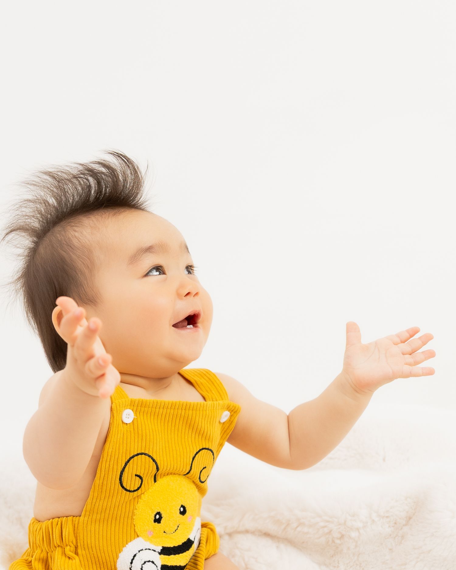 Happy baby in yellow bee romper with wild dark hair, laughing and reaching up with both hands against white background.