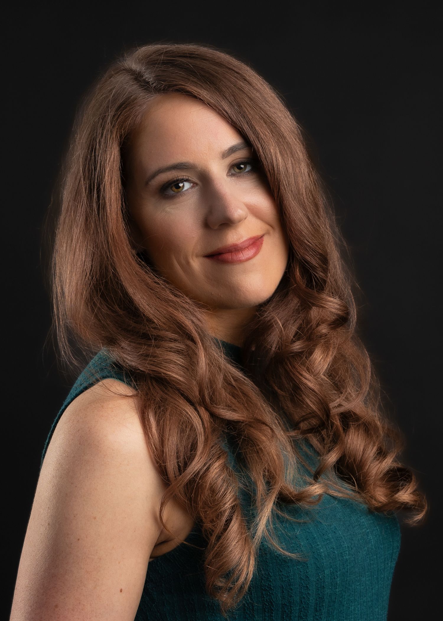 A woman with long auburn wavy hair smiles softly, wearing a teal sleeveless top against a dark background.