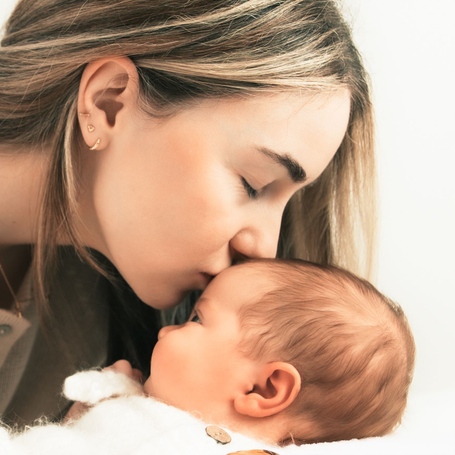 A tender mother gently kisses her sleeping newborn baby on the forehead in a soft, warm close-up portrait.
