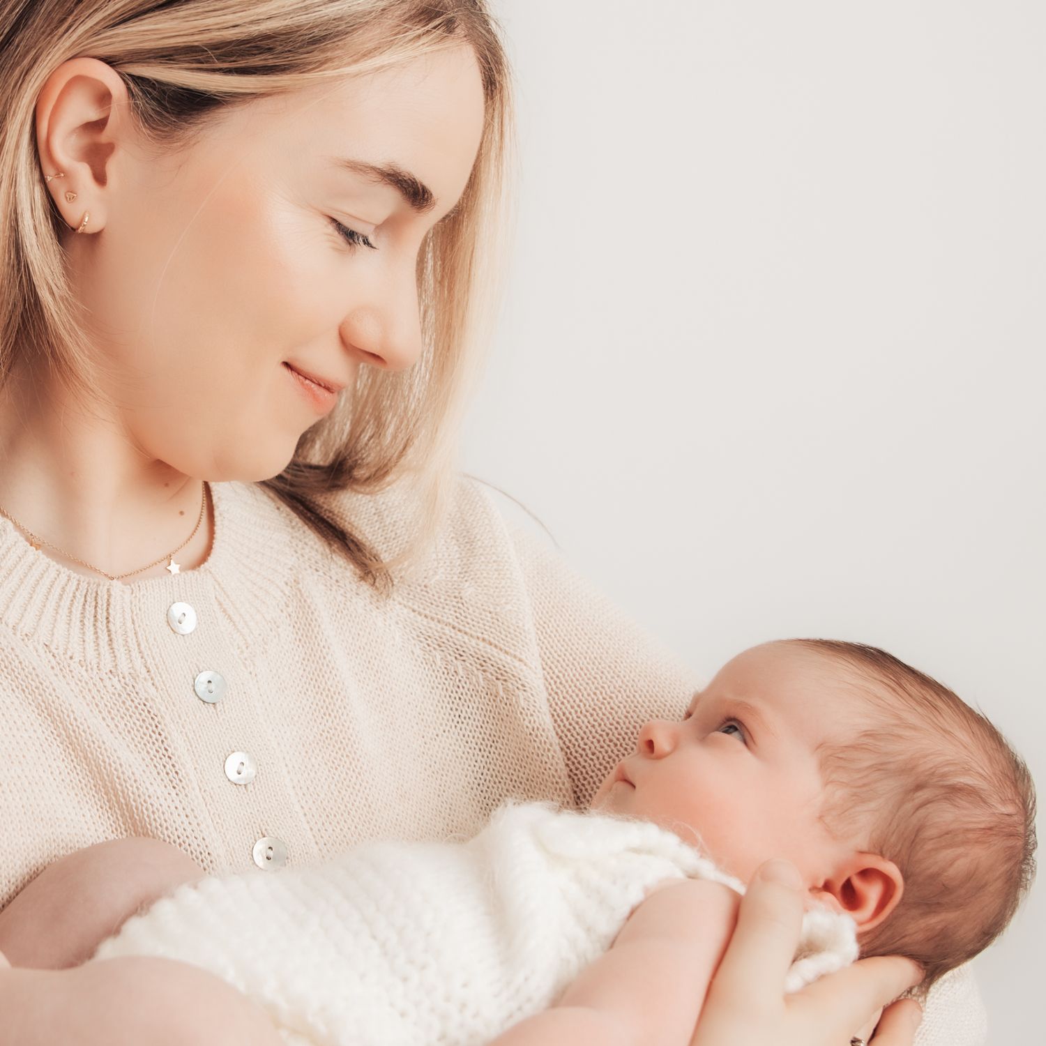 A young mother in a cream sweater tenderly gazes at her newborn baby wrapped in white cloth.