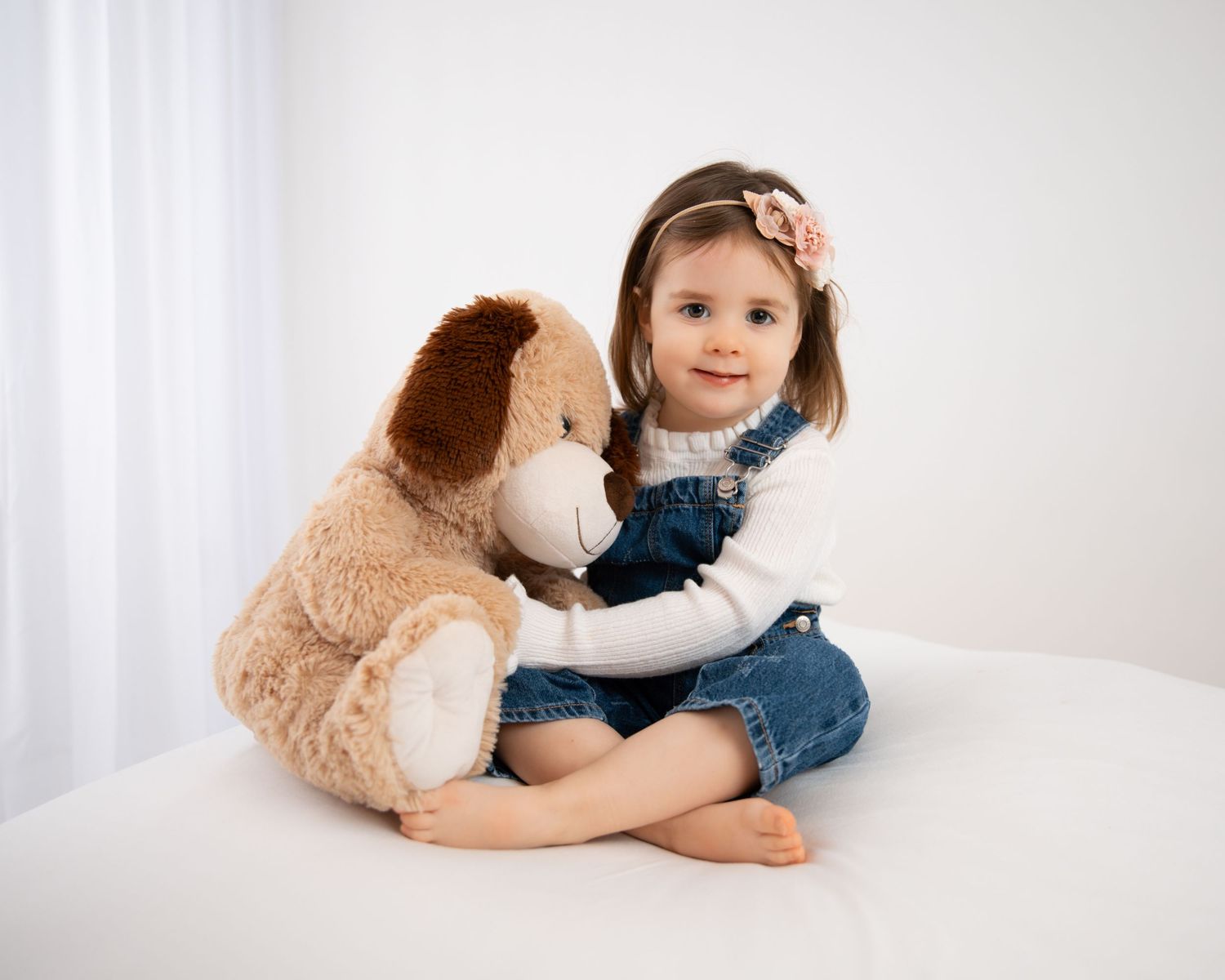 A cute toddler girl in denim overalls sits cross-legged hugging a brown stuffed dog toy on a white background.