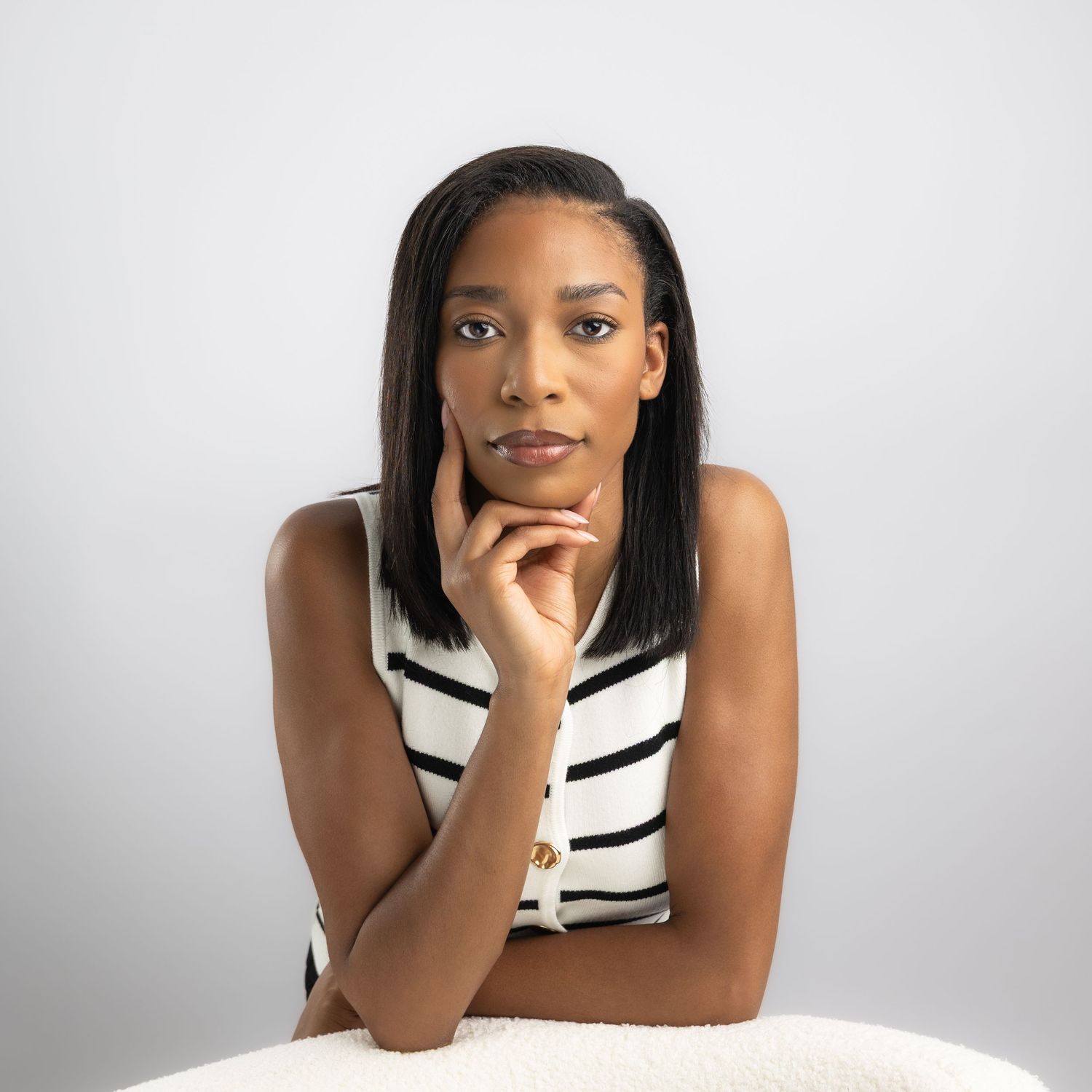 A young Black woman in a striped sleeveless top poses thoughtfully with her chin resting on her hand against a white background.