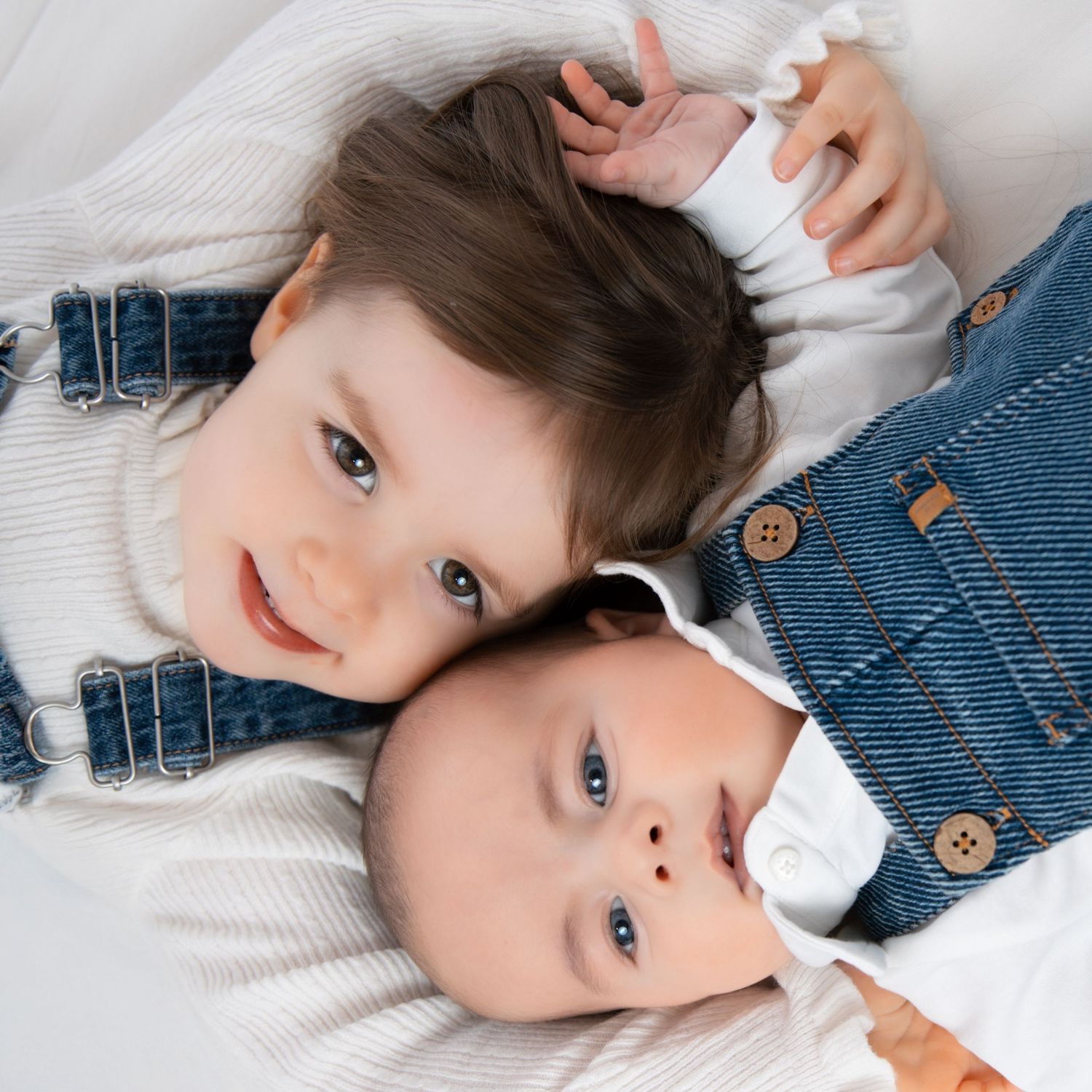Adorable toddler and newborn siblings in denim overalls lying side by side on white background.