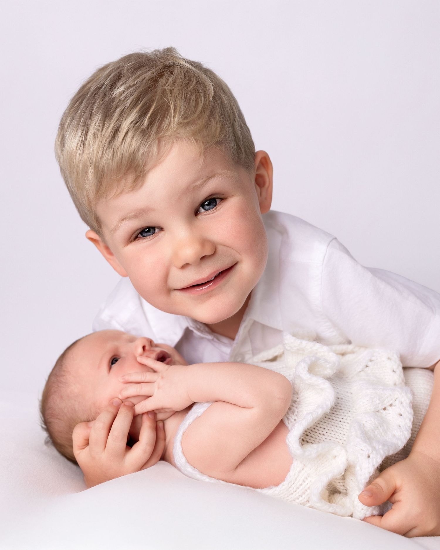Young blonde boy smiling while gently holding a newborn baby in a white studio portrait.
