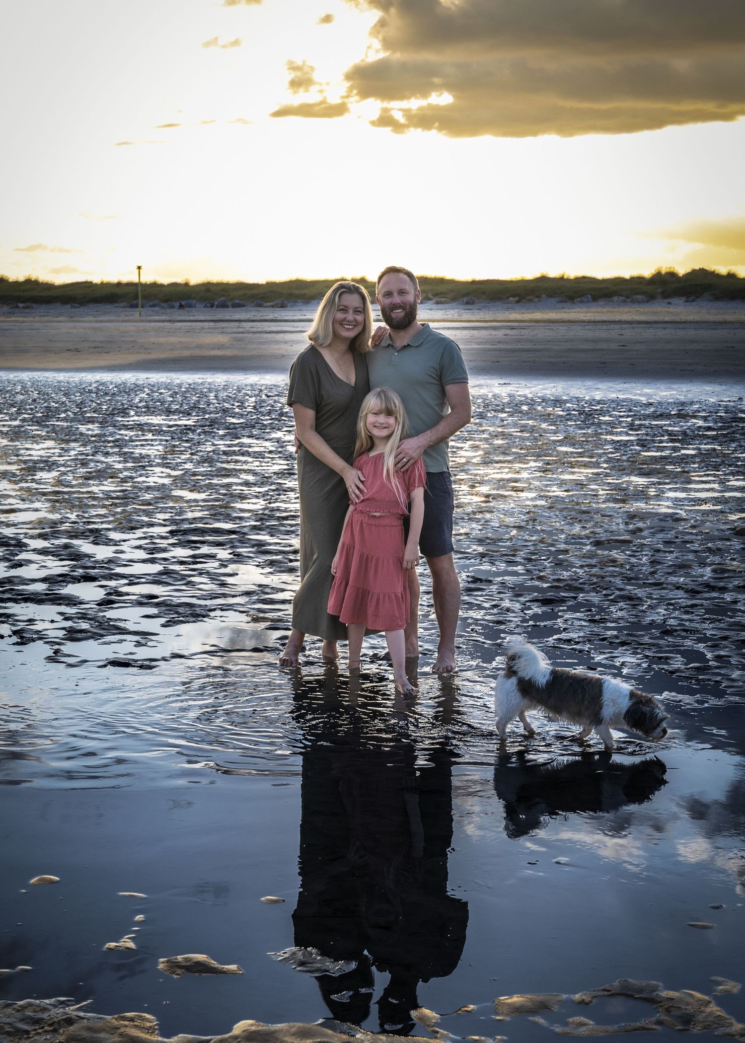 A couple stands in shallow water at sunset creating reflections on the wet sand.