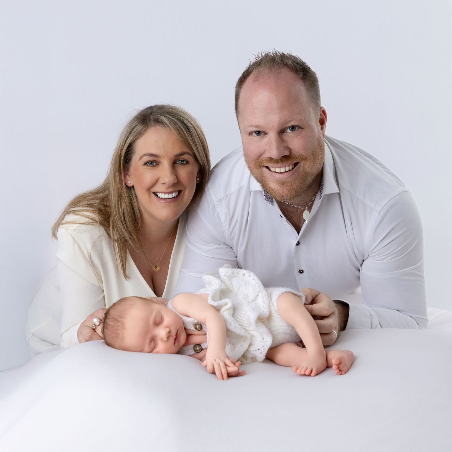 Smiling couple in white outfits posing with their sleeping newborn baby in a professional studio portrait.