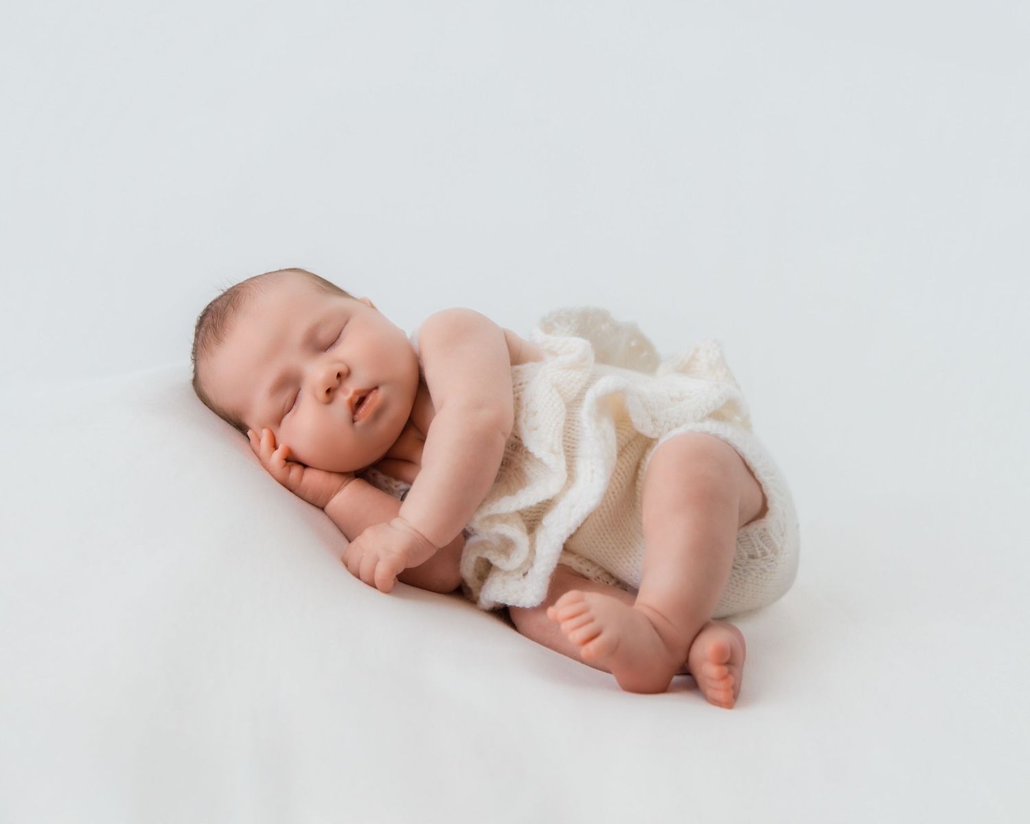 Peaceful newborn baby sleeping curled up on white blanket, wearing cream knit wrap, soft natural light photography.