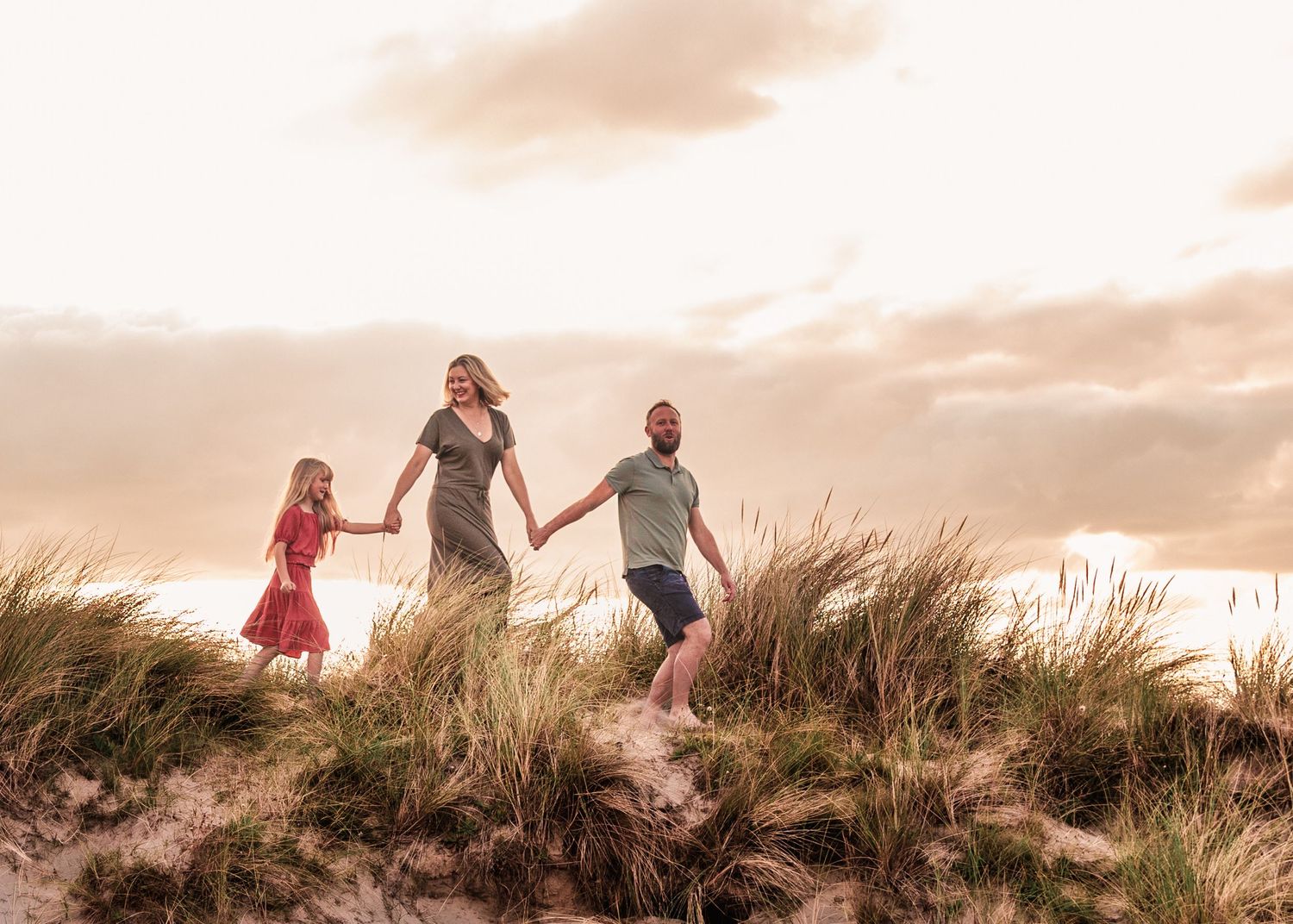 People holding hands while walking along grassy beach ridge at sunset.