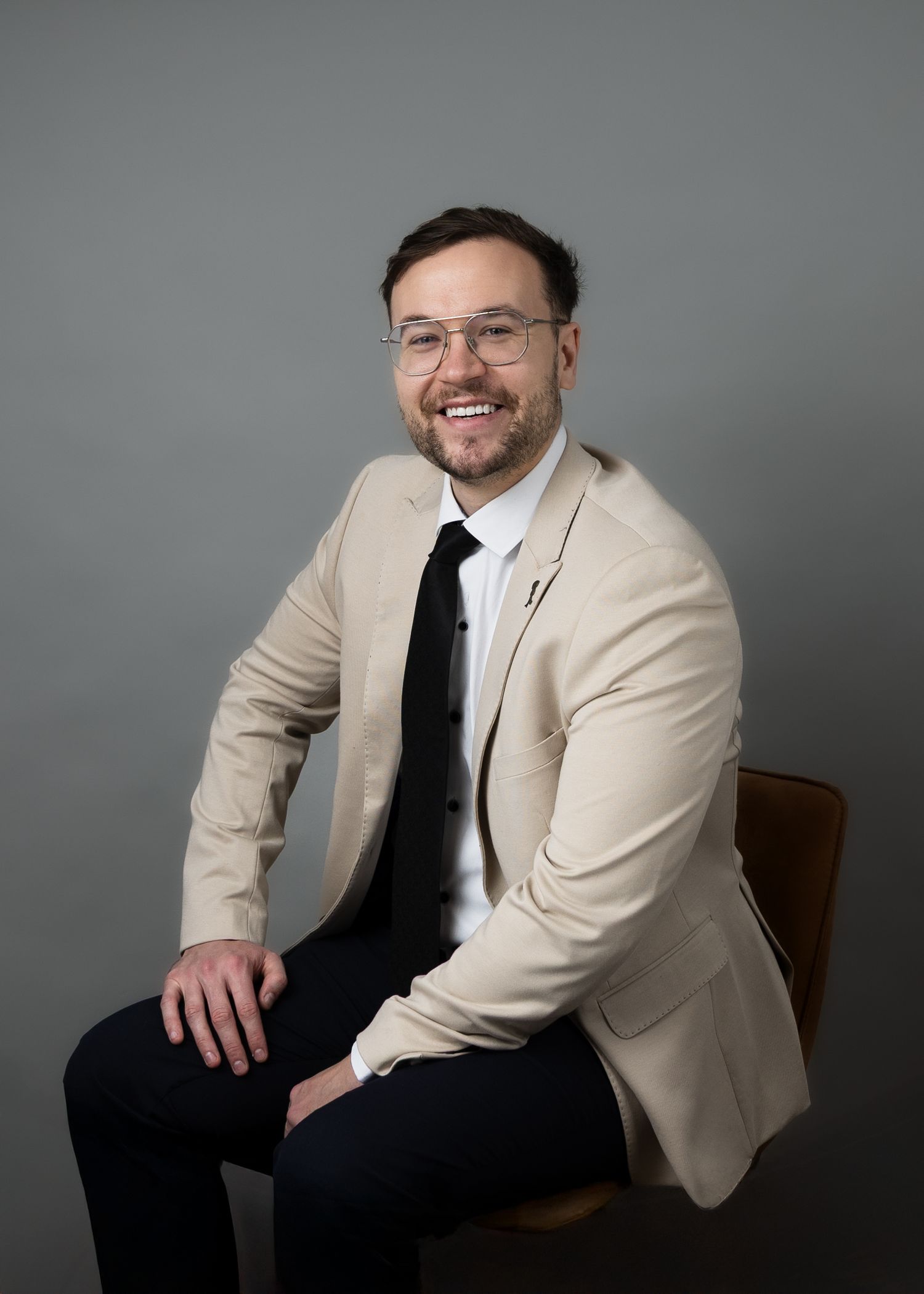 Professional portrait series showing a person in a beige blazer and black tie sitting in different poses against a gray backdrop.