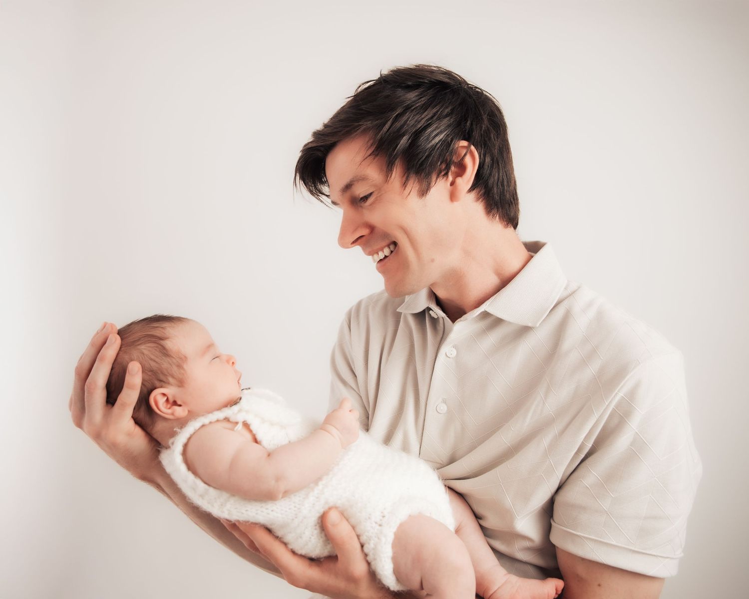 A smiling father gently holds his newborn baby in both hands against a soft white background.