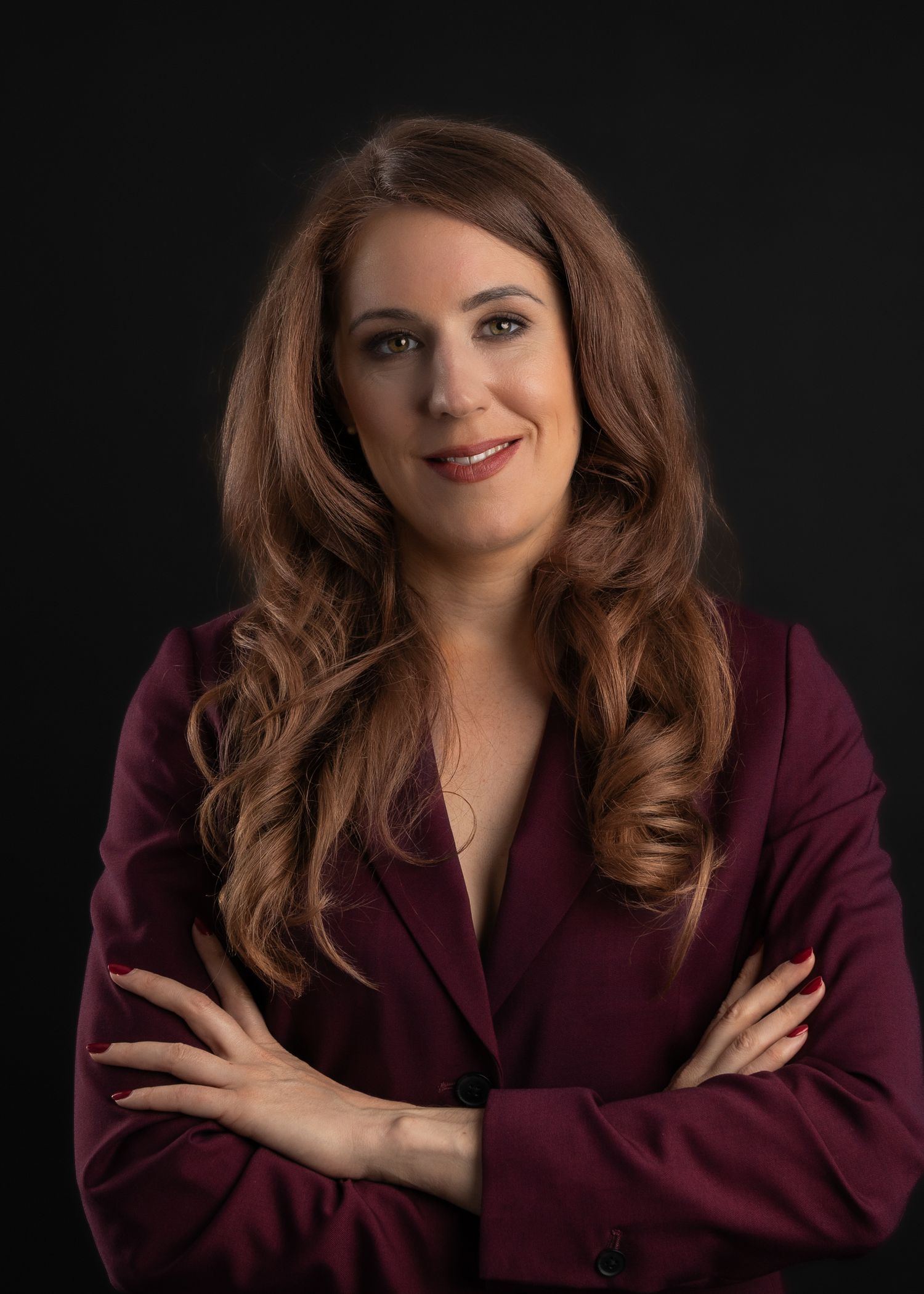 Professional woman with long auburn hair in a burgundy blazer, arms crossed, posing confidently against dark background.