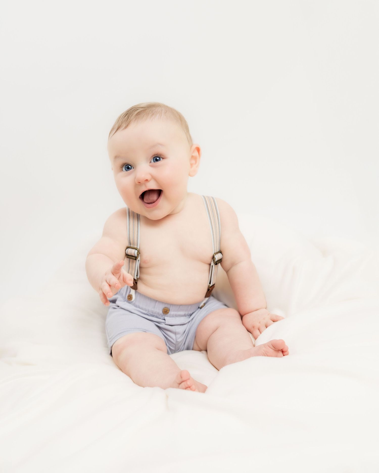 Adorable baby boy sitting up wearing gray shorts and suspenders, smiling with blue eyes on white background.