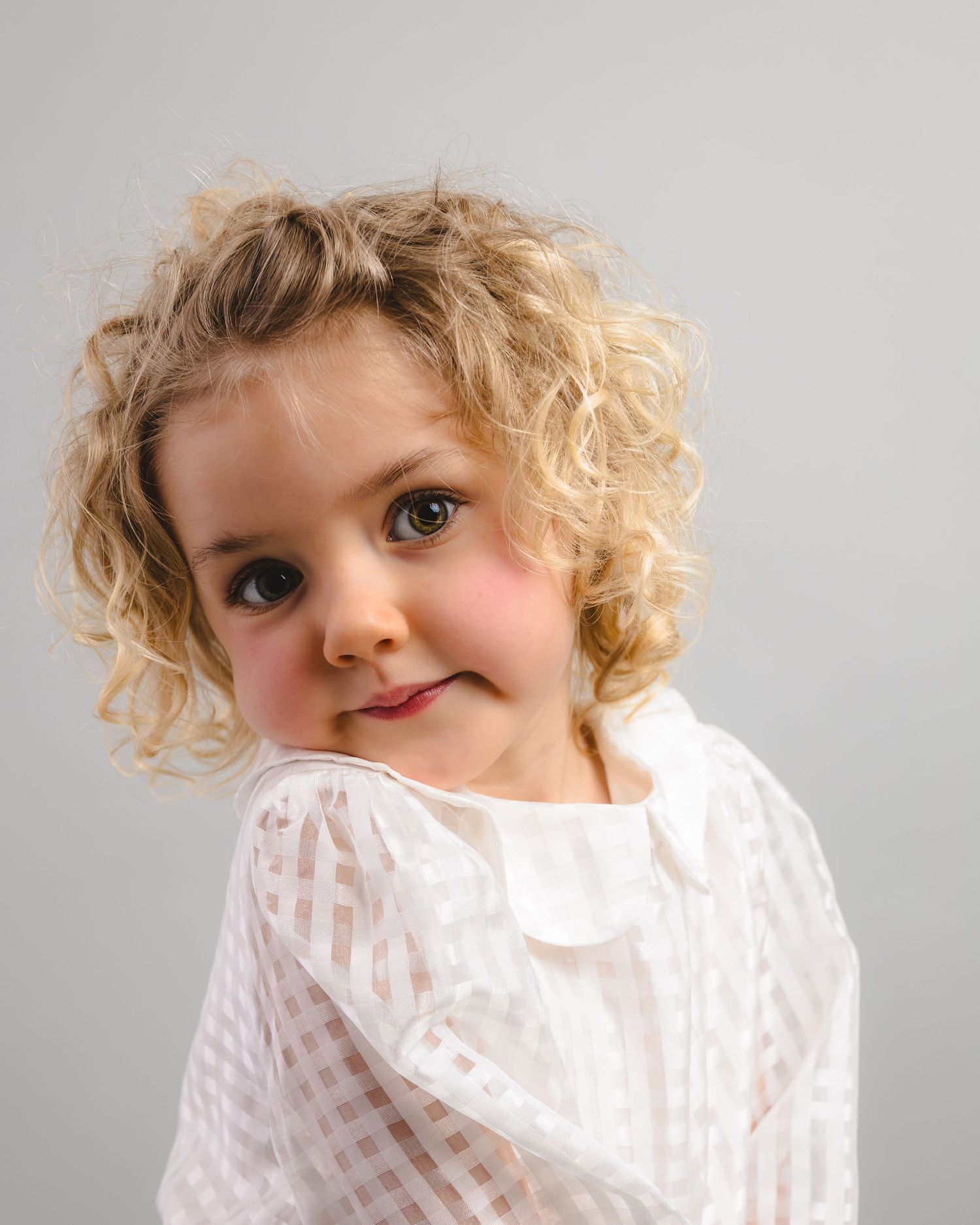 Young girl with curly blonde hair smiles over her shoulder, wearing a white gingham blouse against a grey background.