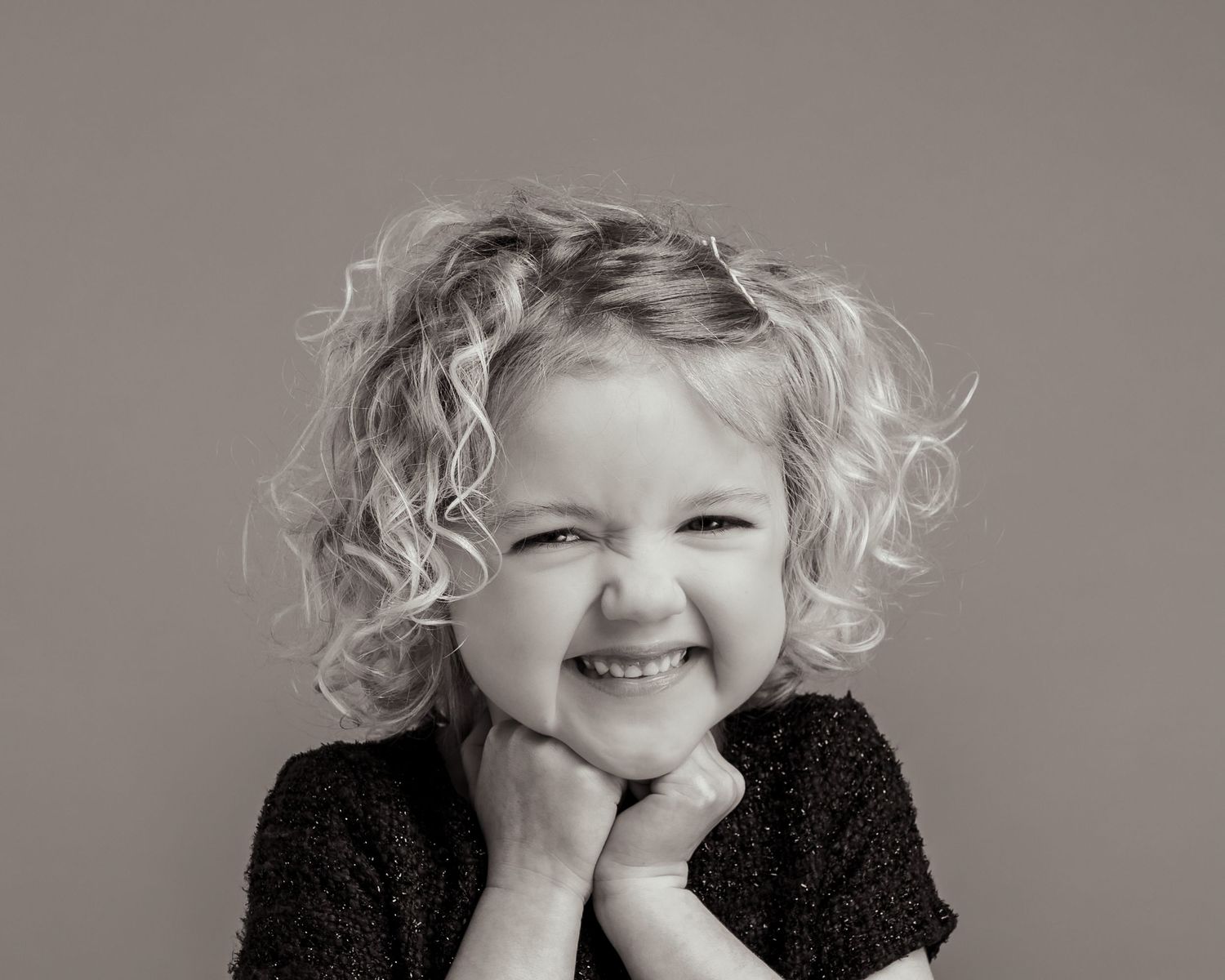 Black and white portrait of a curly-haired toddler girl smiling with hands on chin.