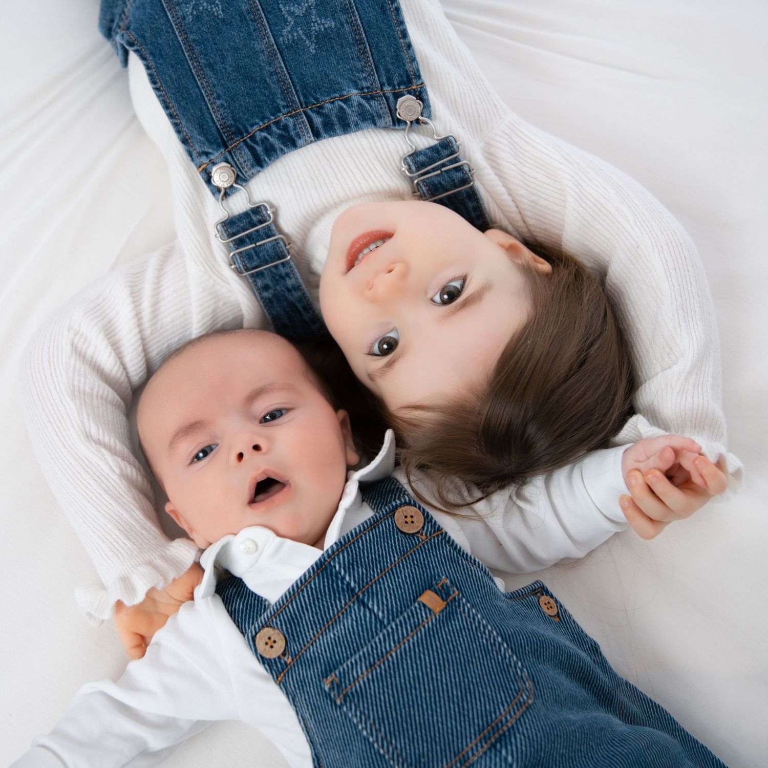 Two young siblings in matching denim overalls lying together, newborn baby and toddler smiling at camera.