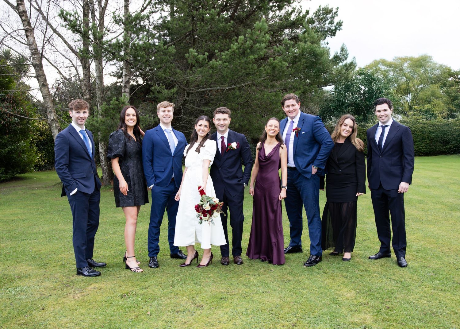 Wedding party members stand in a row on a green lawn for group photos during an outdoor celebration.
