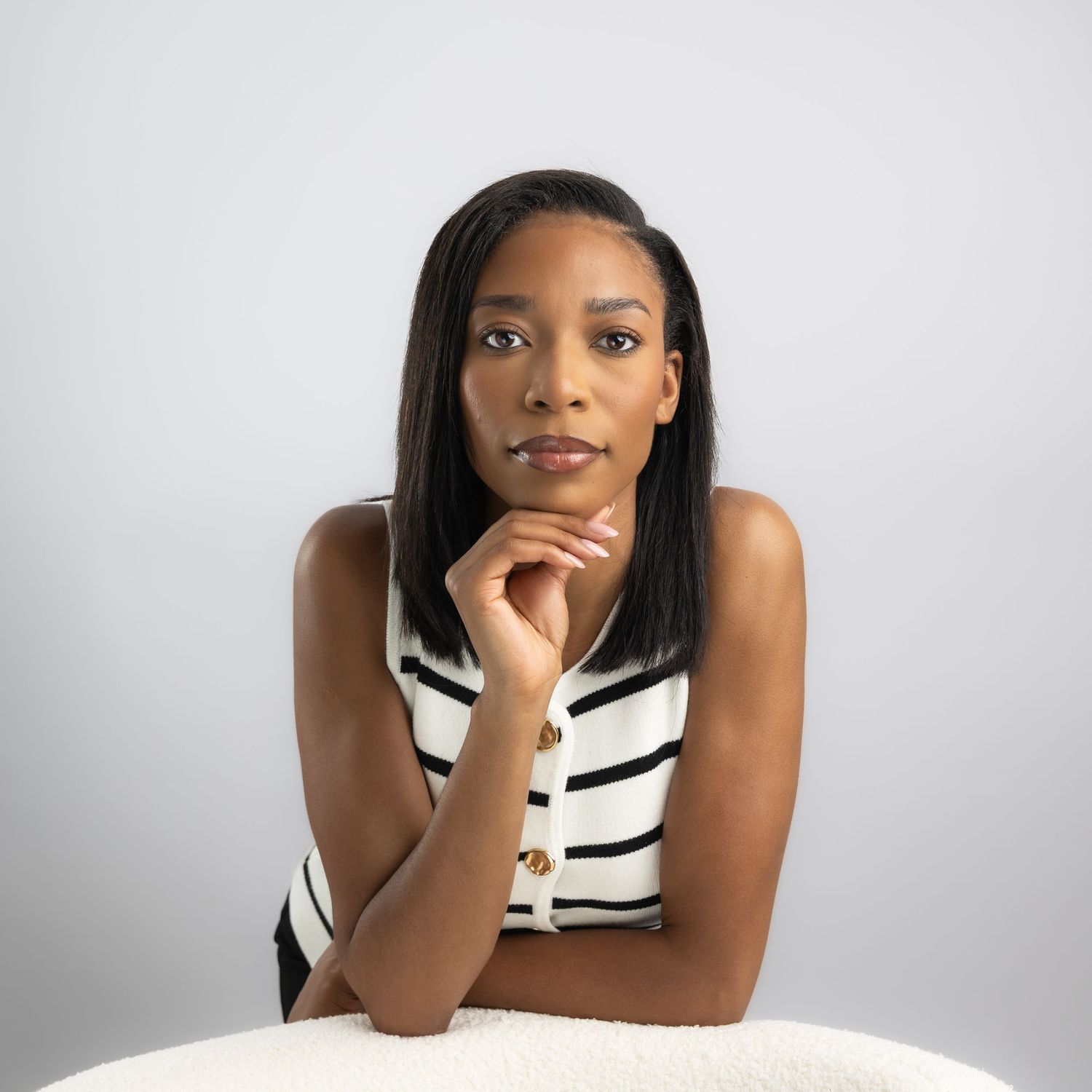 A young Black woman in a striped sleeveless top poses thoughtfully with her chin resting on her hand against a white background.