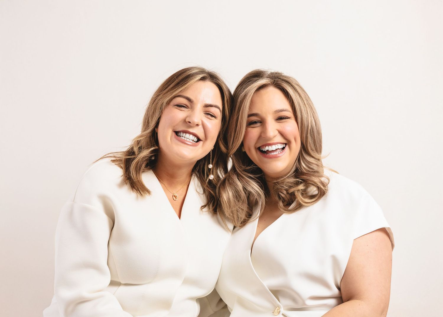 Two smiling women in white outfits posing together against a light background in a professional photo.
