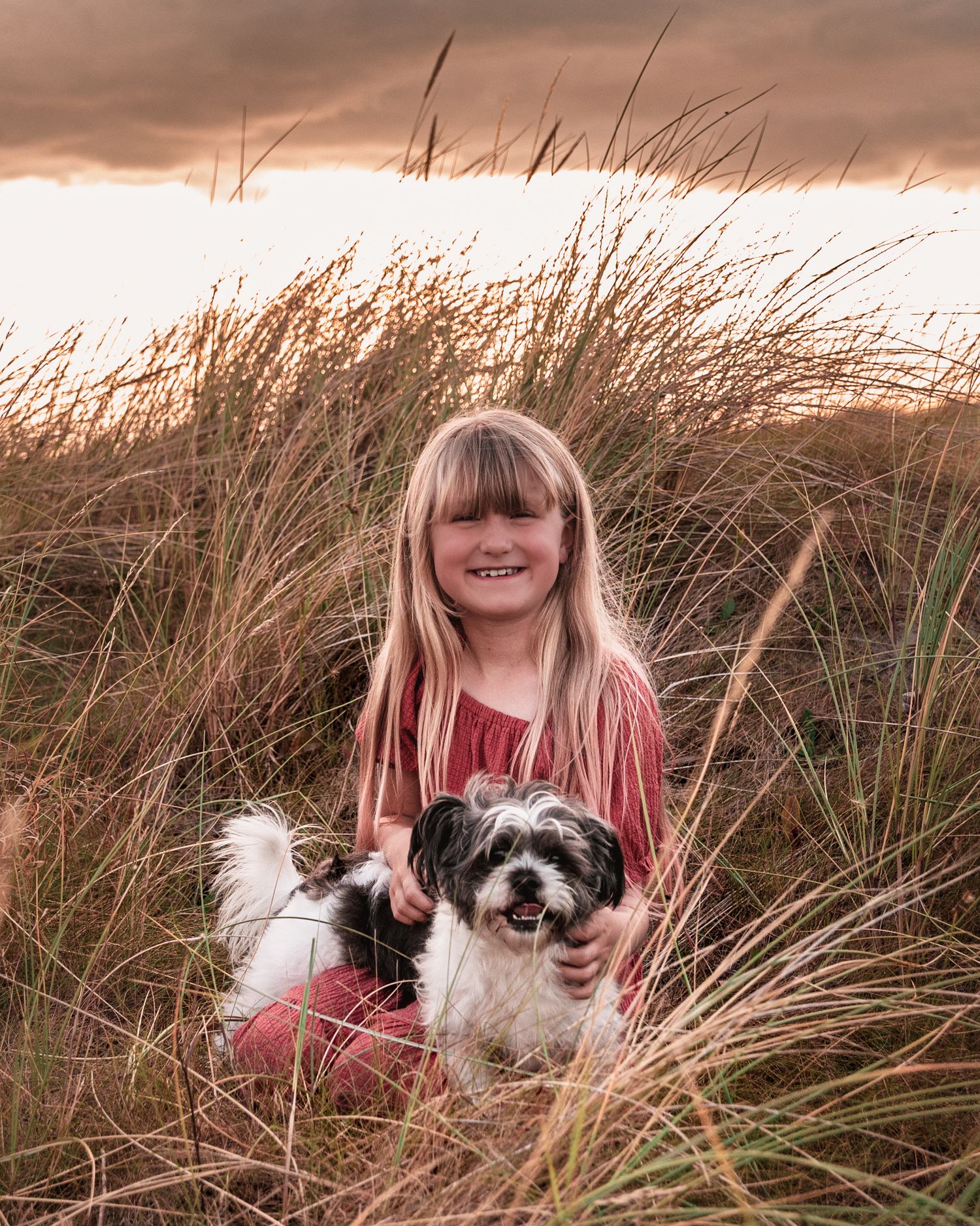 A child and small dog sitting together in tall beach grass during sunset.