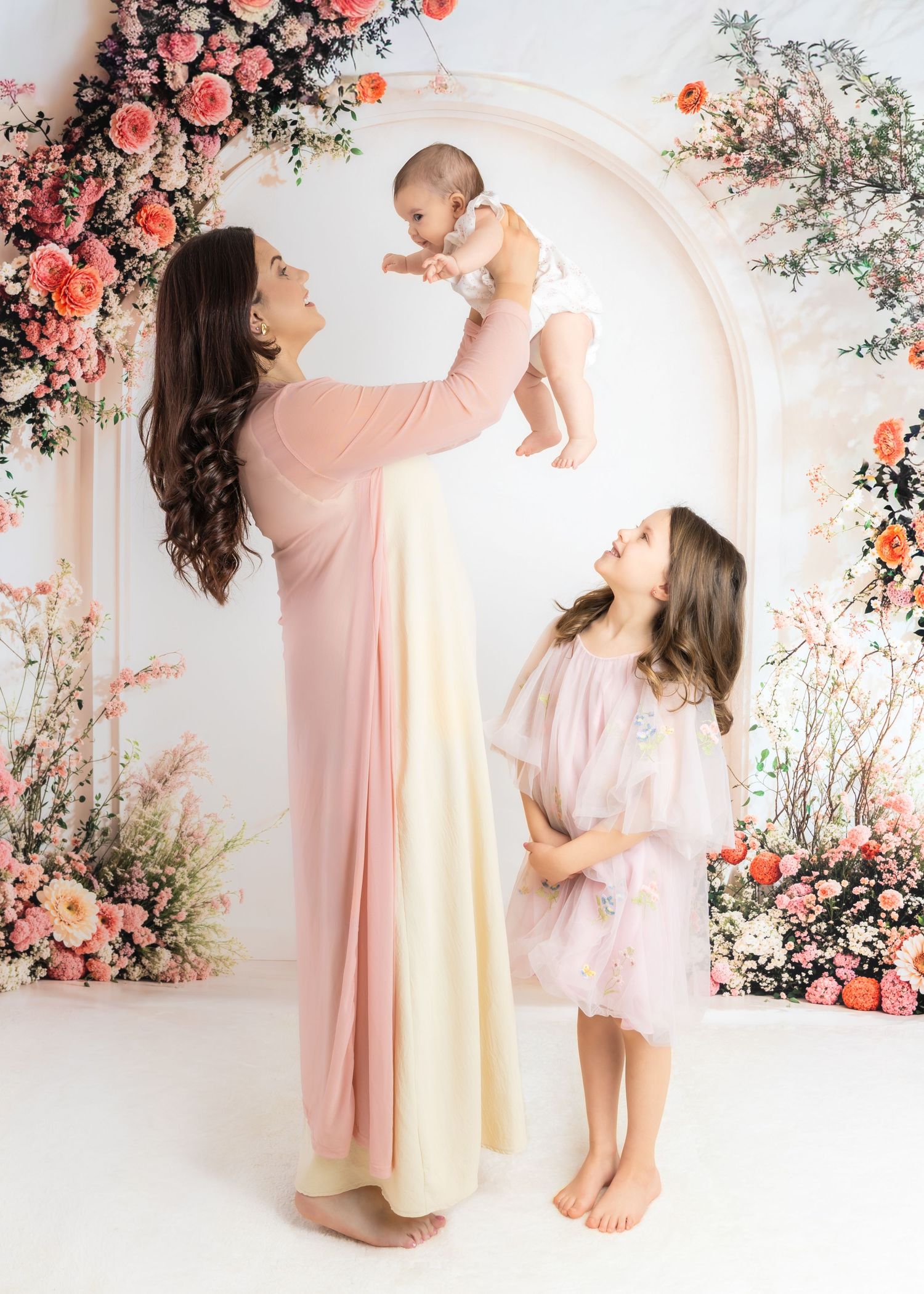 Ethereal portrait session in pink featuring mother and baby against floral arch backdrop with coral roses and greenery.