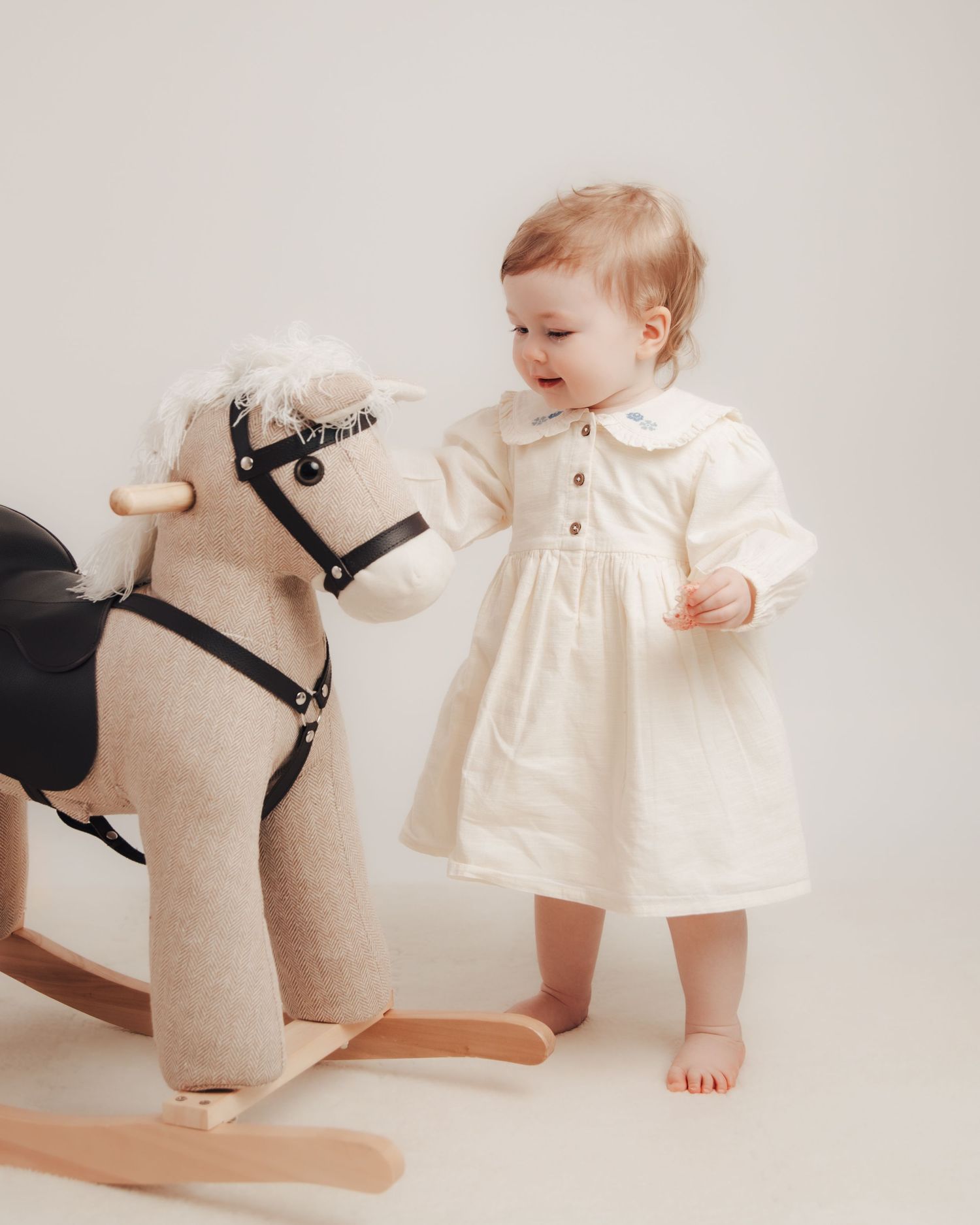 A toddler in a white dress stands barefoot next to a rocking horse toy against a neutral background.