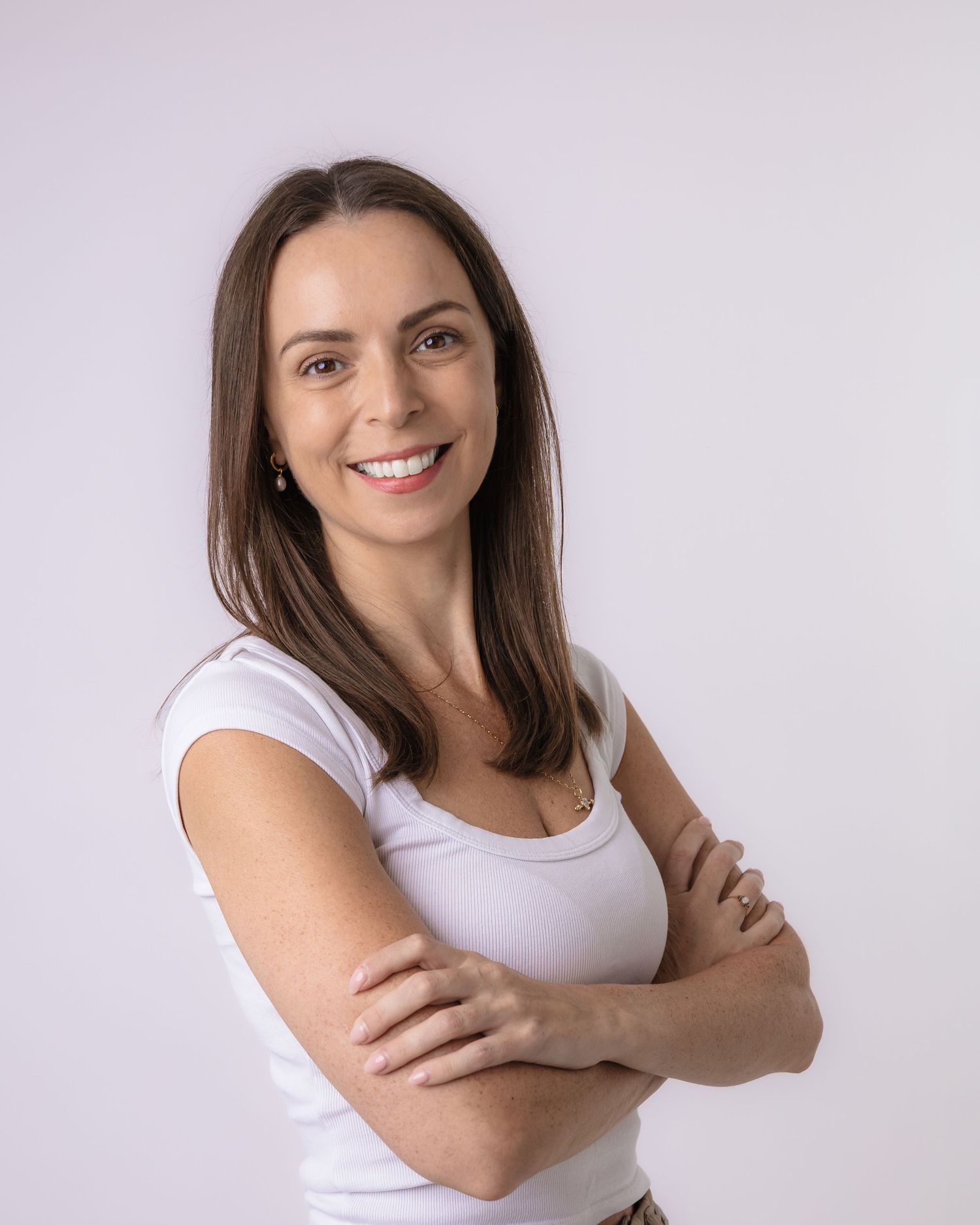 Smiling woman with brown hair in white t-shirt, arms crossed, posing confidently against light background.
