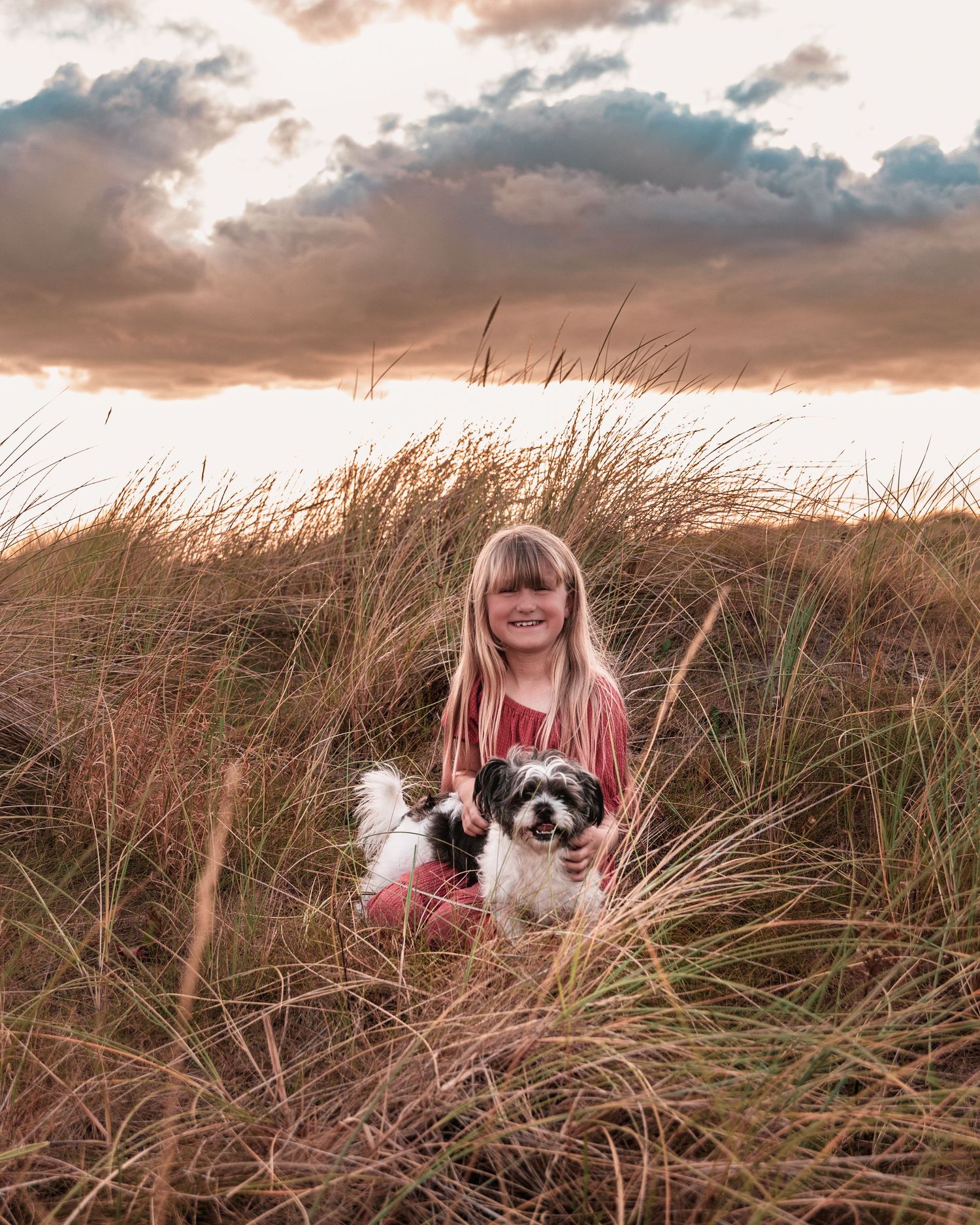 Child cuddling with small dog in beach grass during sunset.
