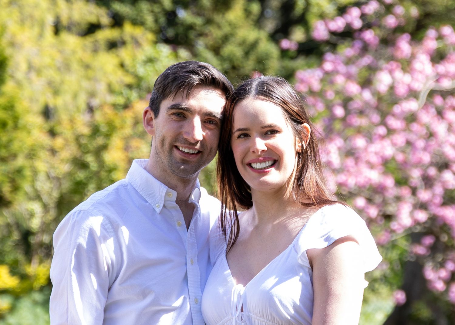 A couple poses outdoors in white clothing with pink cherry blossoms and yellow foliage in the background.