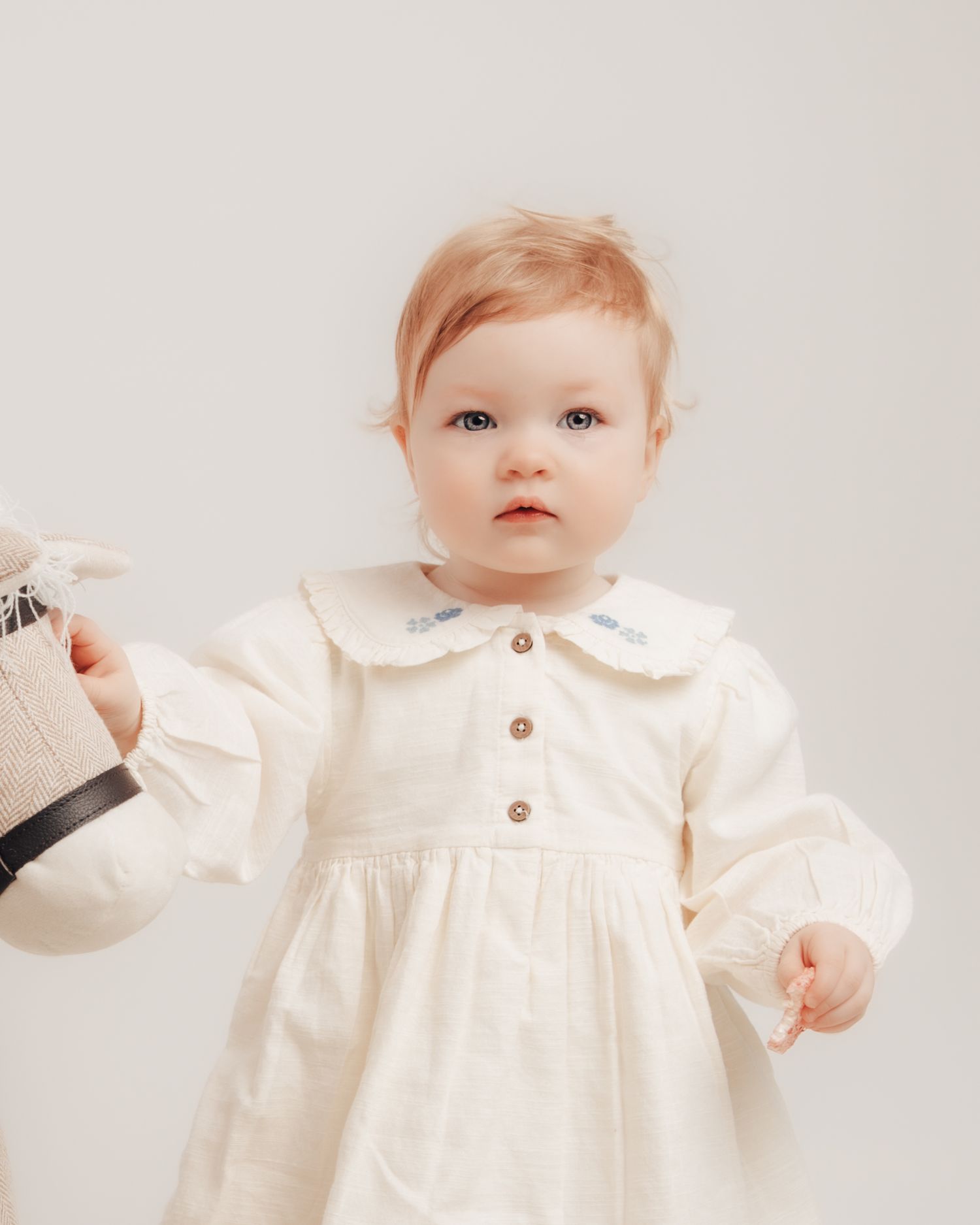 Baby girl in cream dress with peter pan collar and blue embroidery, holding a toy.