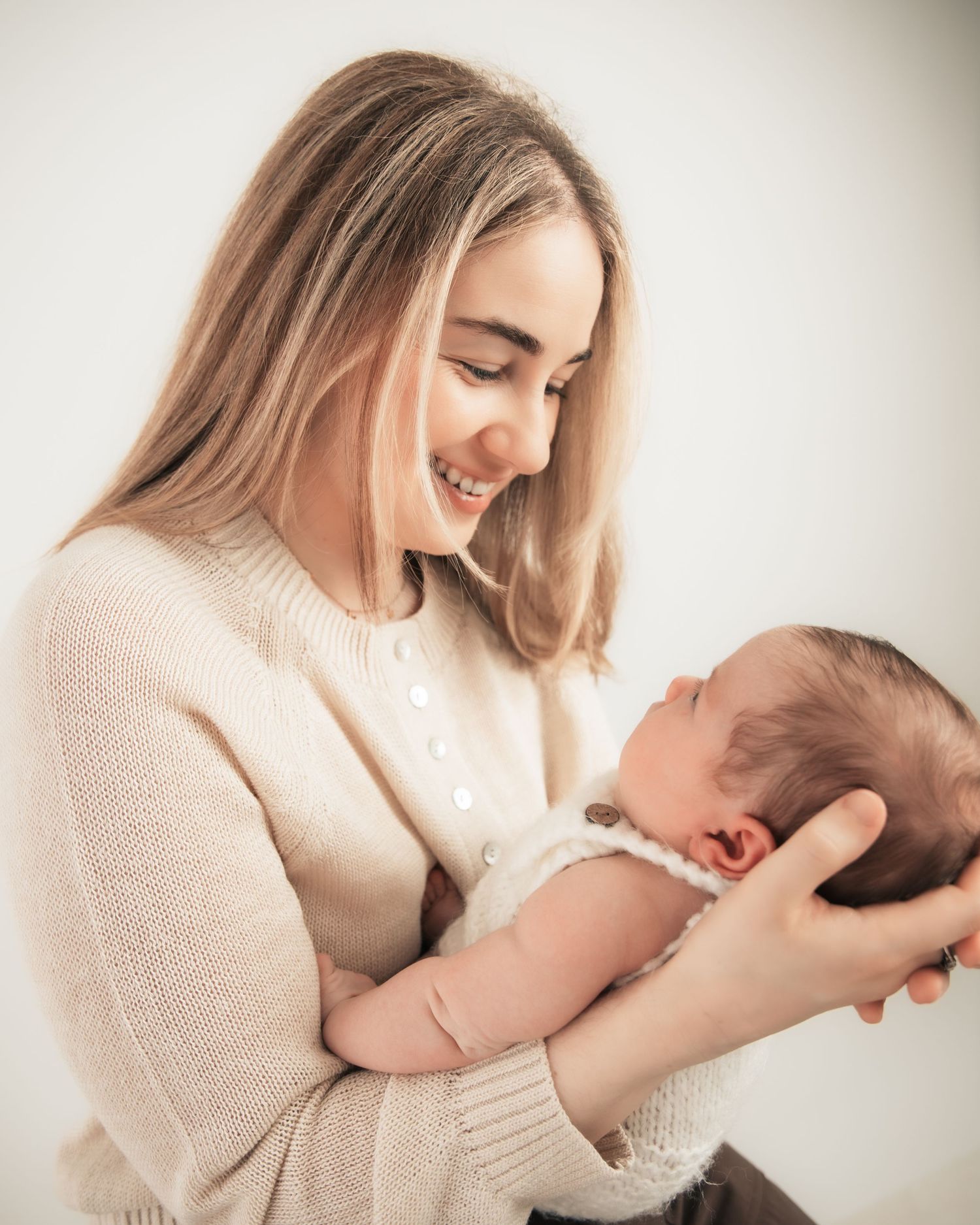 Young woman with long blonde hair smiling warmly while cradling a newborn baby in her arms.