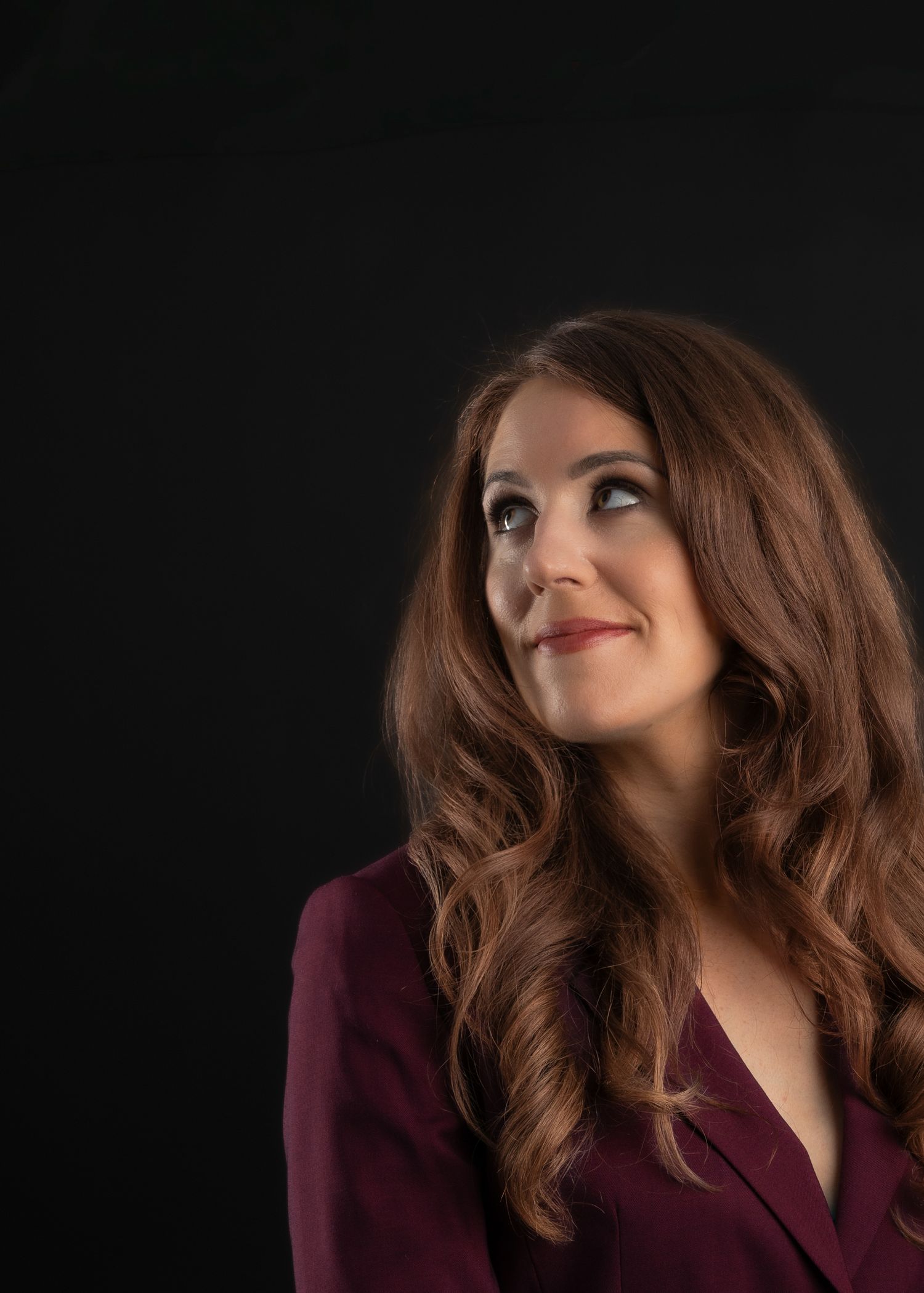 A woman with long auburn hair in a burgundy blazer gazes upward against a dark studio background.