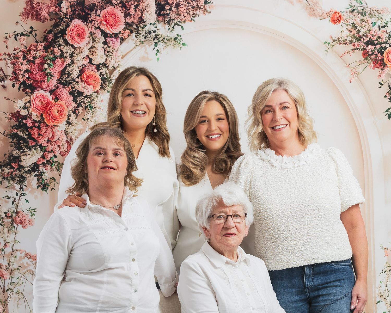 A group poses together in front of a white wall decorated with pink and coral floral arrangements.