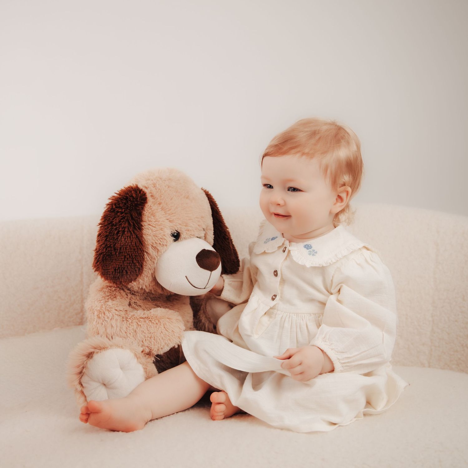 A smiling baby in a cream dress sits beside a large plush puppy stuffed animal on a white chair.