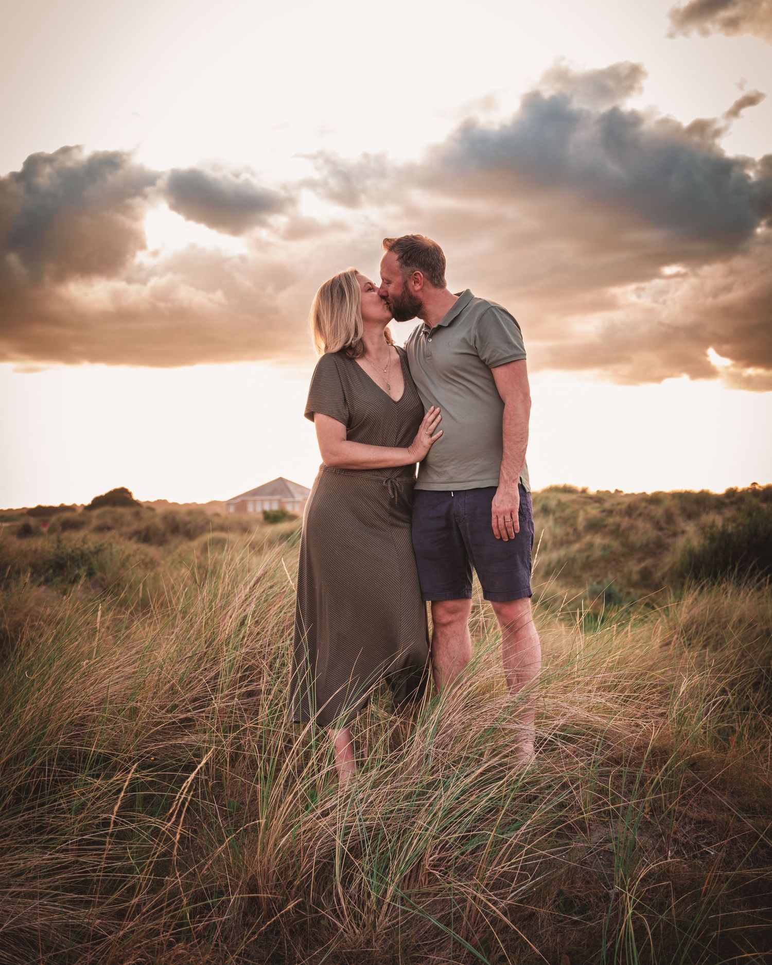 Couple embracing in tall grass during a dramatic sunset photoshoot on the beach.