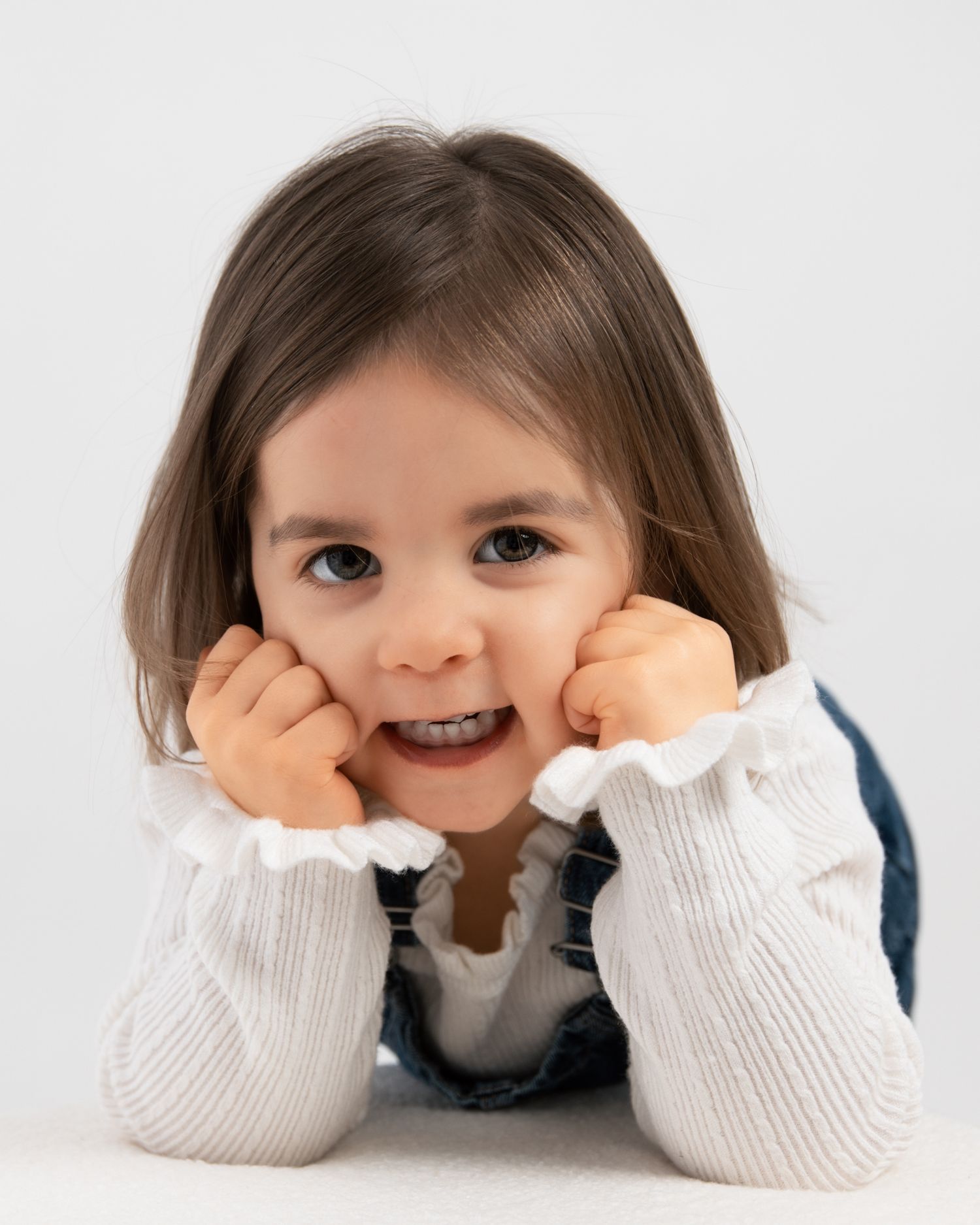 Cute young girl resting chin on hands, smiling at camera, wearing white sweater and denim overalls.