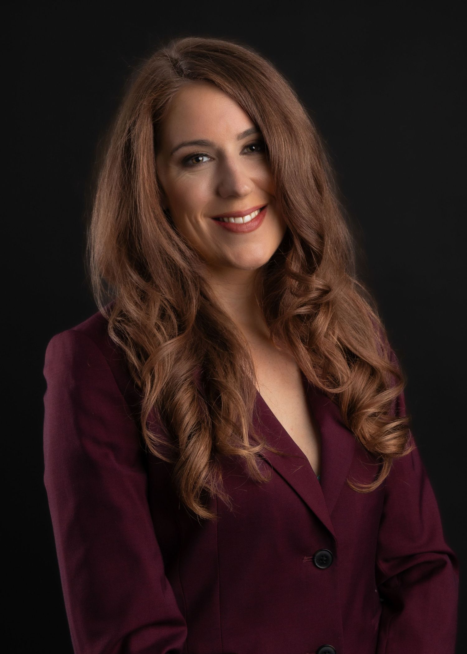 Professional headshot of a smiling woman with long auburn hair wearing a burgundy blazer against a dark background.
