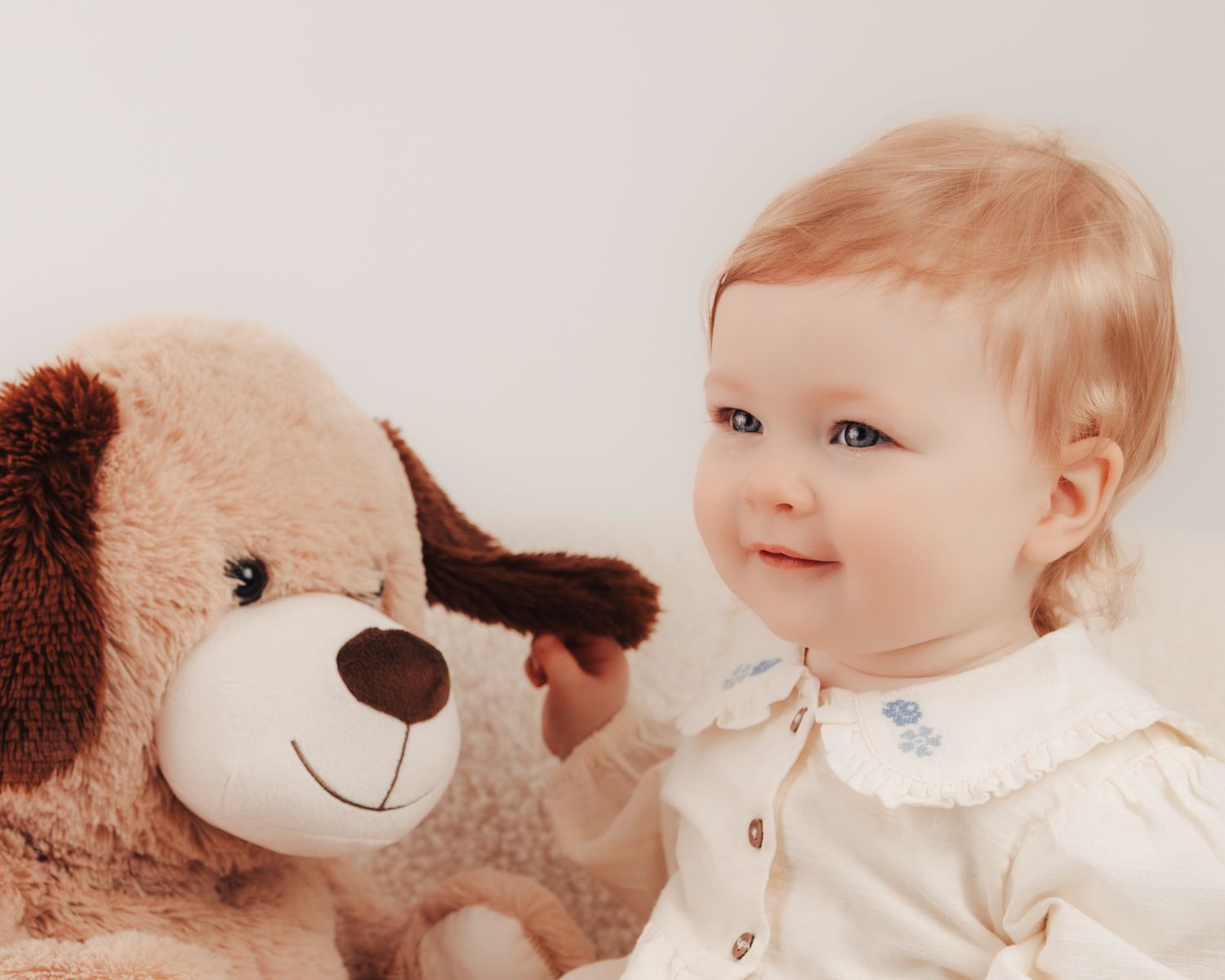 A smiling baby with blue eyes in a white outfit sits next to a brown teddy bear against a light background.