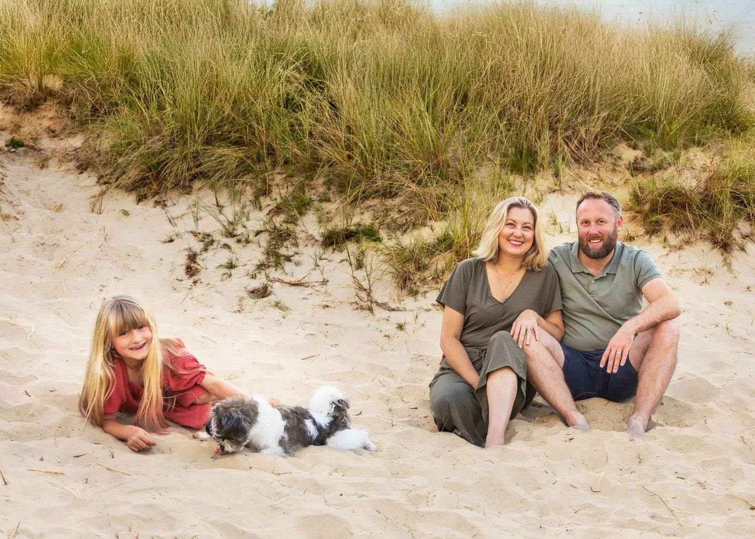 Family members lounging on sandy beach near grass-covered dunes.