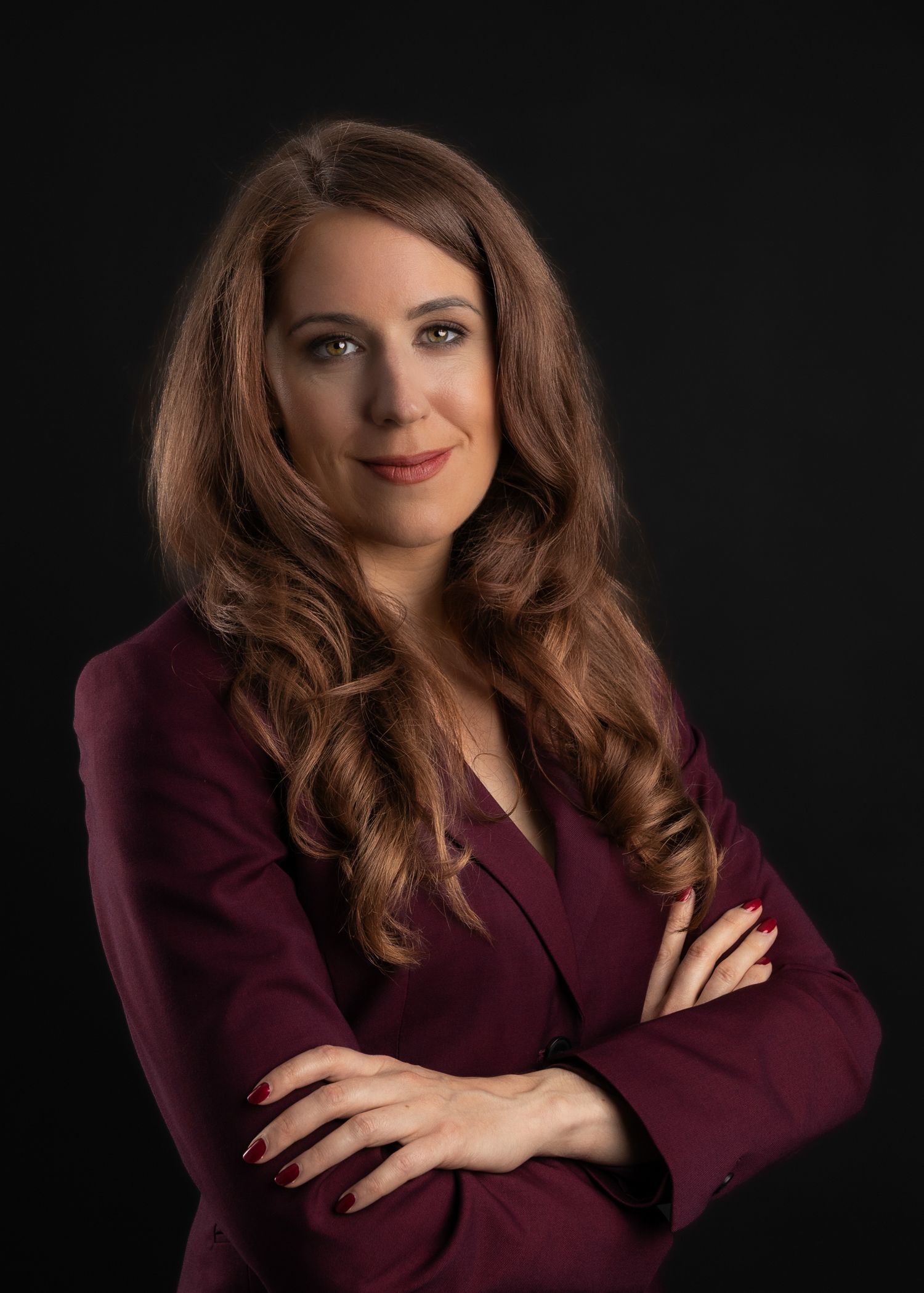 Professional woman in burgundy blazer with crossed arms, posing confidently against dark background.
