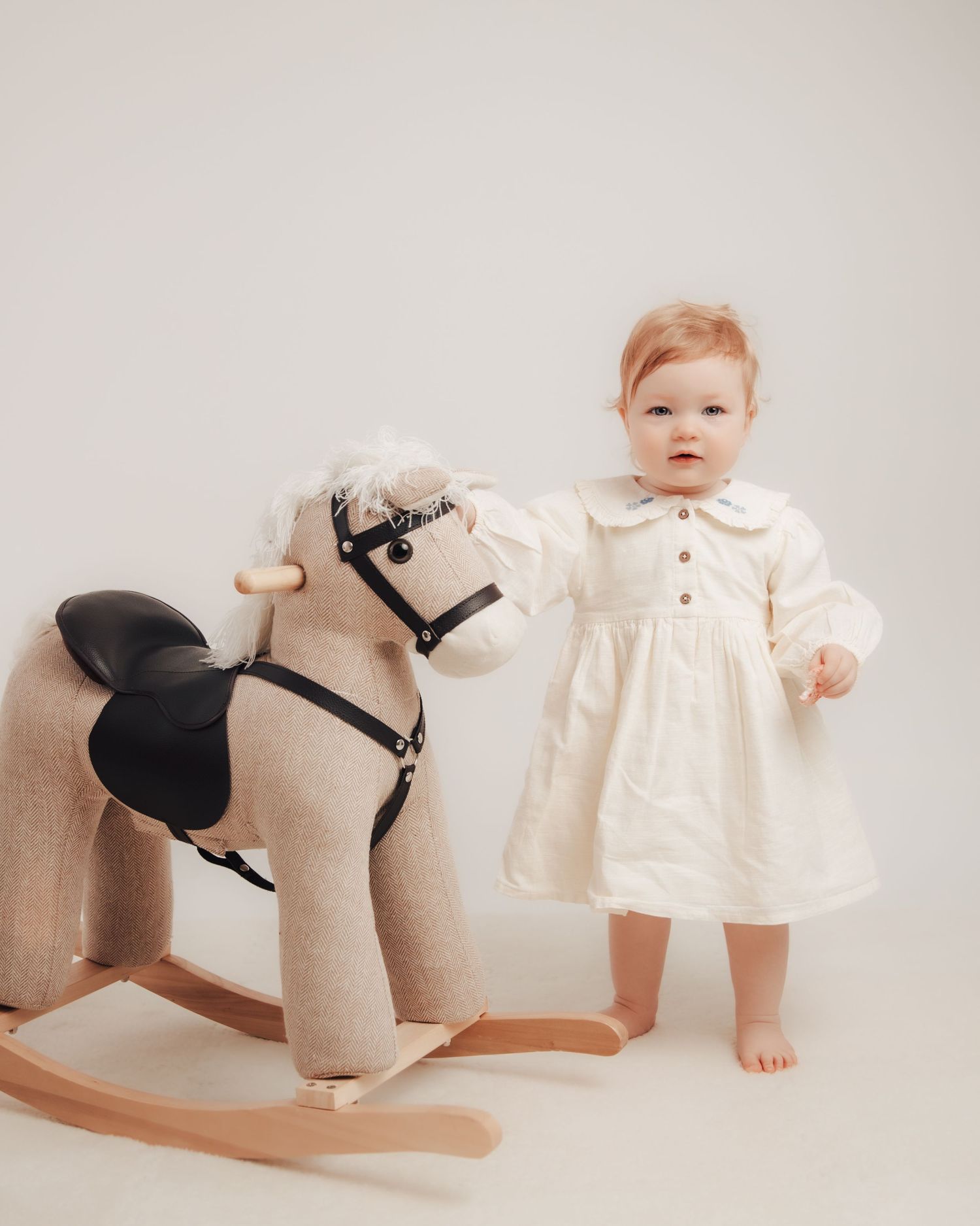 Toddler girl in cream dress standing next to a rocking horse toy.