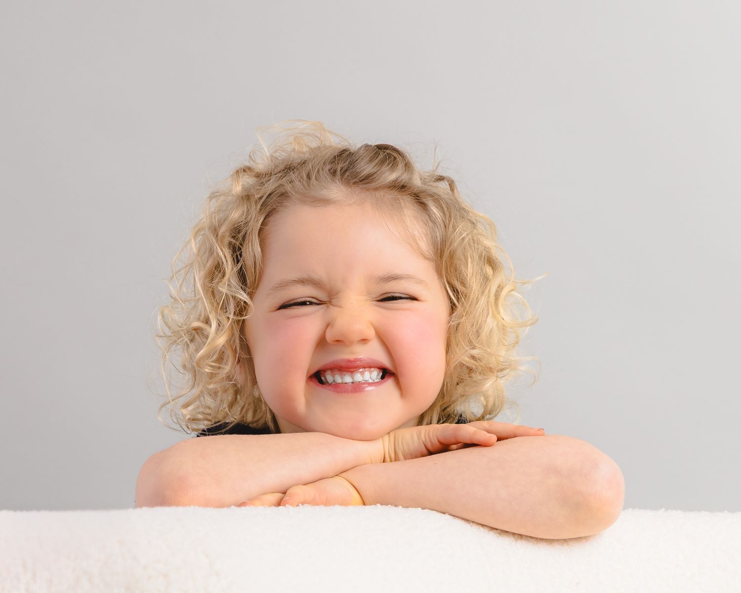 Happy toddler with curly blonde hair resting chin on arms, smiling brightly against a light gray background.