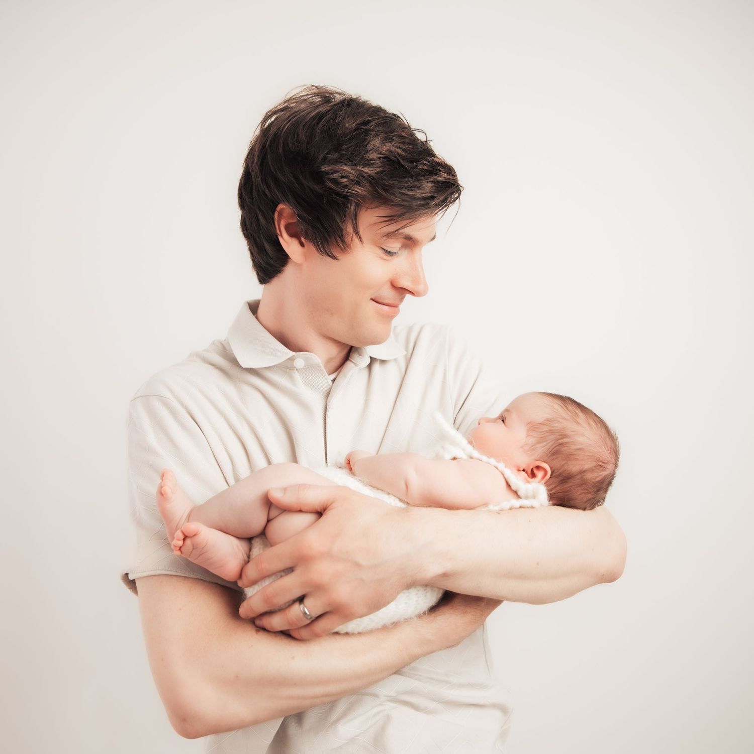 A young father smiles lovingly while cradling his newborn baby against a soft white background.