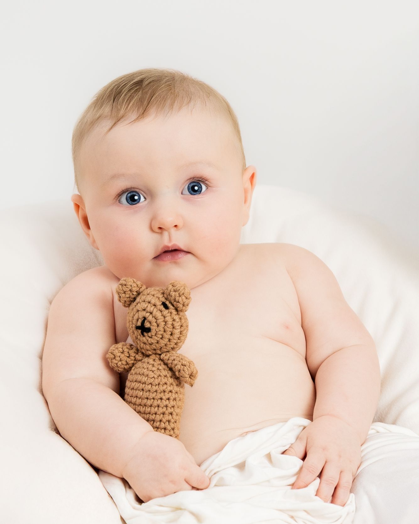 Baby lying on white bedding with a small brown crocheted teddy bear companion.