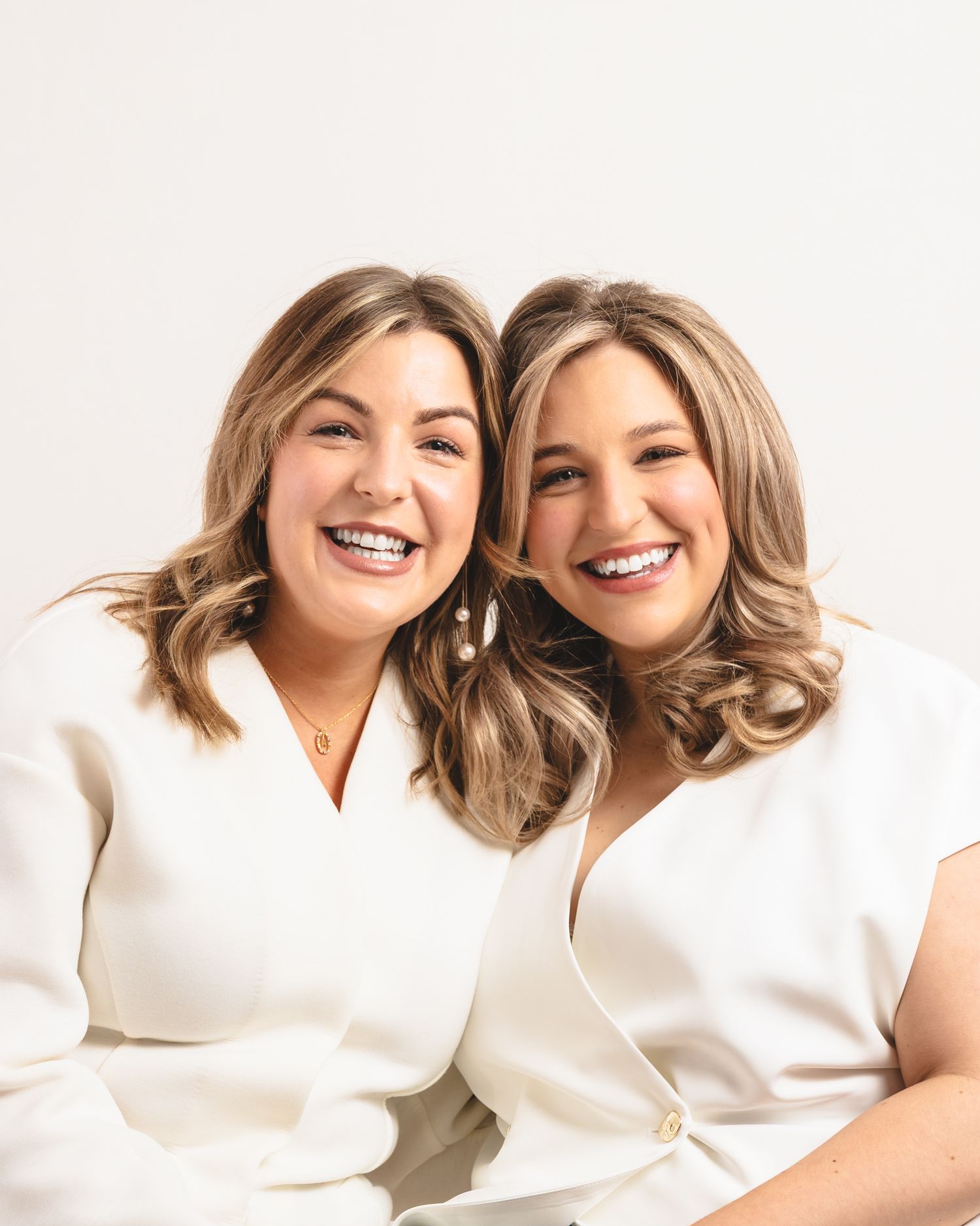 Two women in white outfits sit together smiling warmly against a light background.