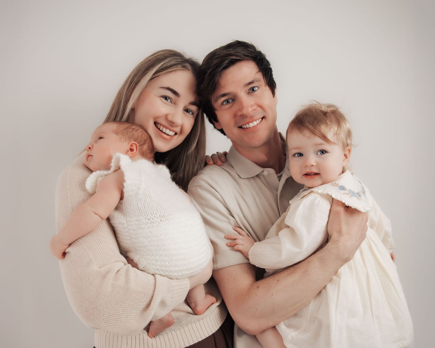 Young couple smiling and holding two babies, a newborn and a toddler, posing together for a family portrait.