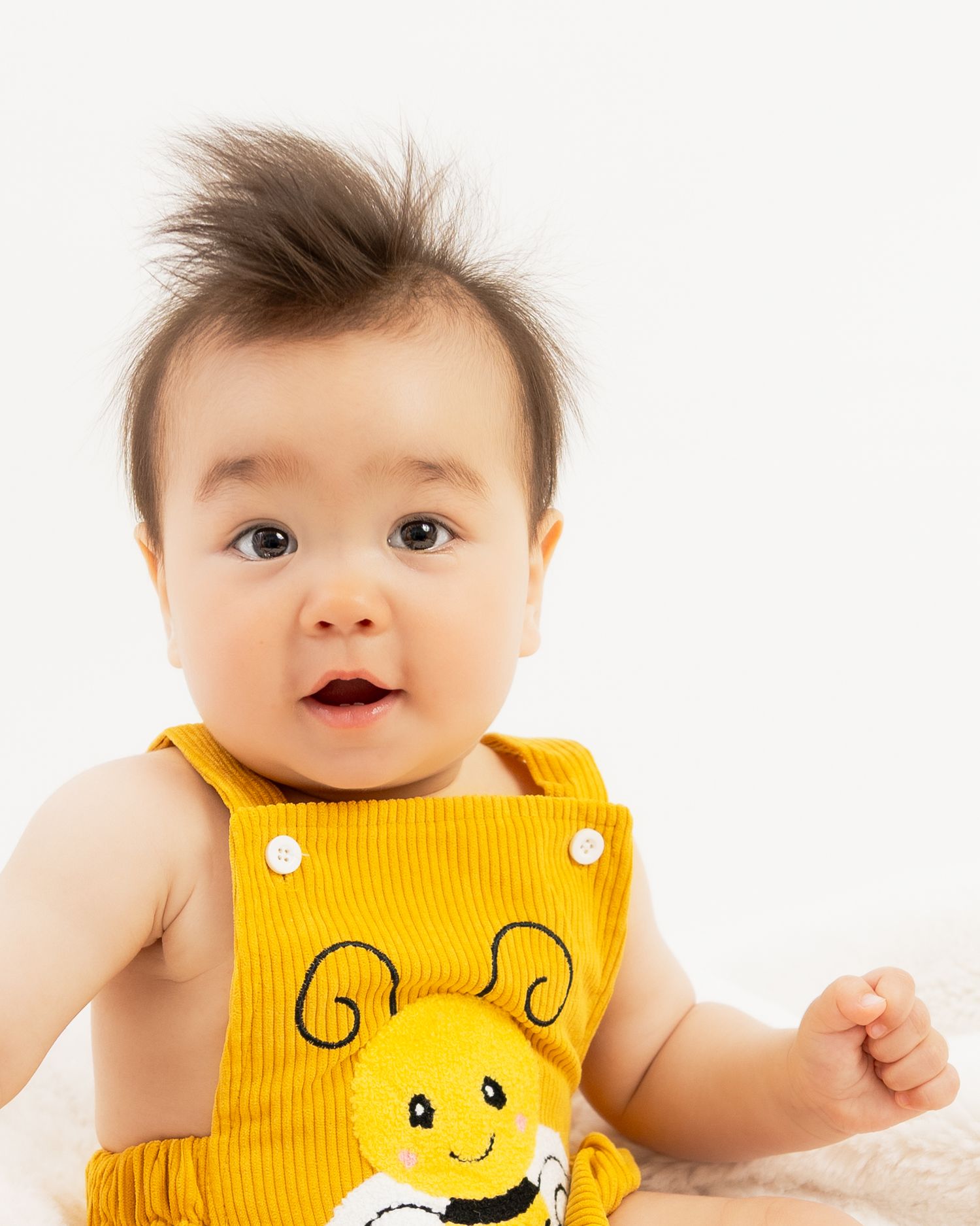 Adorable baby in yellow bee overalls with wild dark hair, wide eyes, and open mouth, posed against white background.